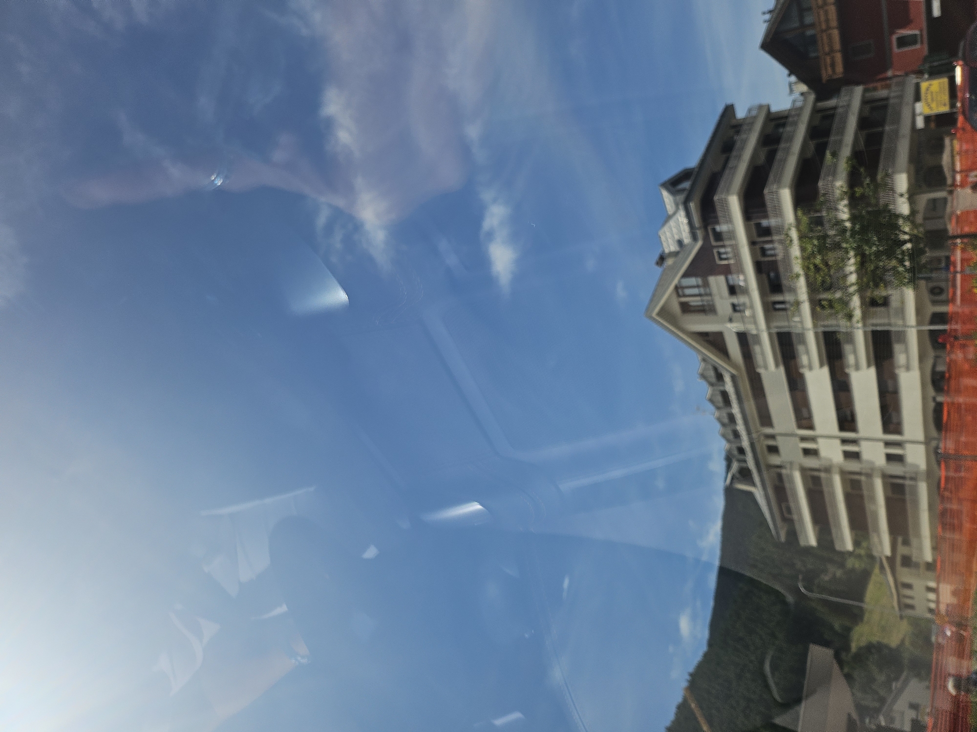 The image is a photo taken from a vehicle, likely a car, looking out at a bright blue sky with wispy clouds. There are reflections on the windshield, showing parts of the vehicle's interior, including a person's shoulder and head on the left side of the frame, and the top of what appears to be a dashboard or car frame in the upper portion of the image.
In the mid-ground, a multi-story apartment building with balconies and a pitched roof is visible. The building is light-colored with dark window frames. To the left of this building, a lush green mountain slope covered in trees rises sharply. Further to the right, a smaller, dark red building with wooden balconies is partially visible.
In the foreground, an orange construction fence is present, with a hint of a person in blue clothing to the far left. A yellow sign is also visible near the base of the apartment building. The overall scene suggests a daytime view of a town nestled against a mountainous backdrop, likely in the Italian Alps given the mention of Aprica, Italy.