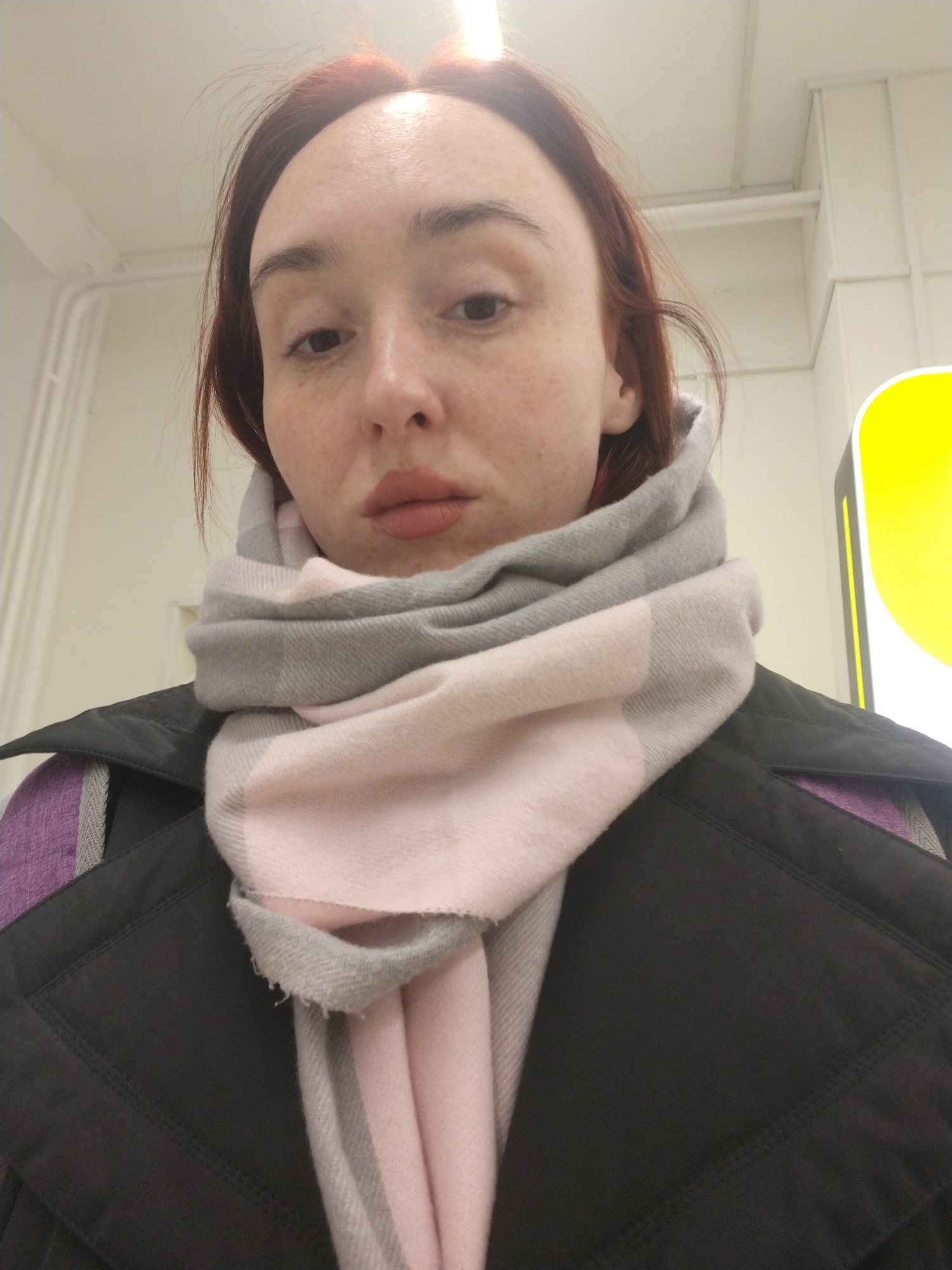 A woman with red hair is standing indoors, wearing a black jacket and a gray and pink scarf. The background shows a white ceiling with pipes and a yellow sign. The location is Izhevsk, Russia.