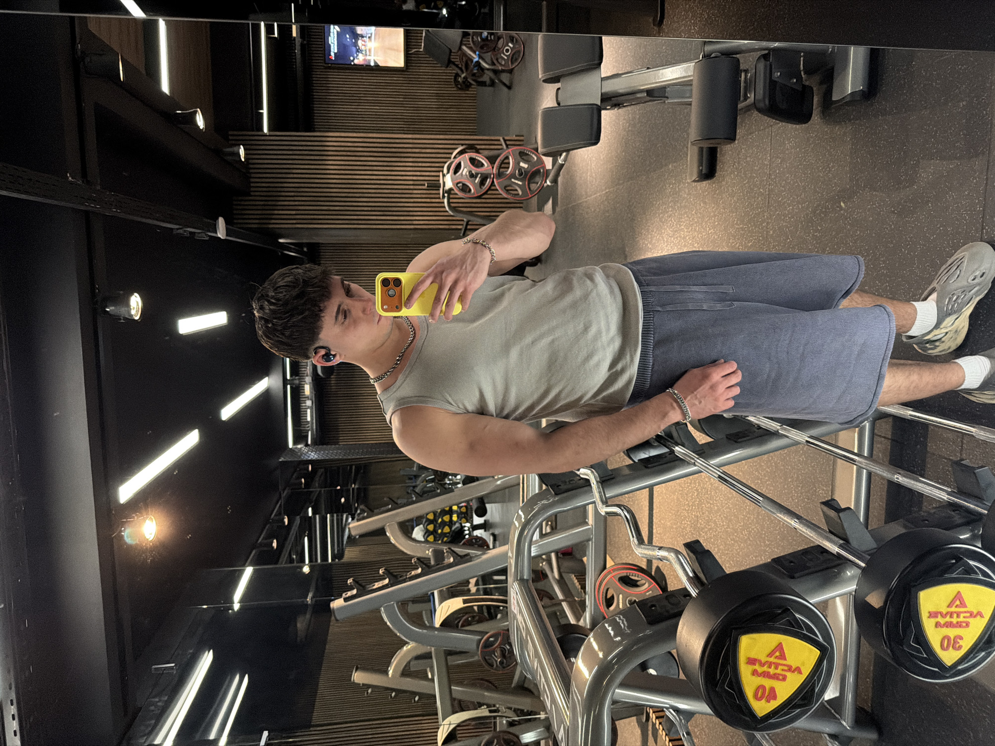 The scene depicts a male individual taking a mirror selfie inside an Evita Fitness & Spa gym facility in Sofia, Bulgaria. The subject, a male with short, dark hair, wears a dark earbud in his left ear, a silver chain necklace, a light grey ribbed tank top, dark grey shorts with a drawstring, white socks, and grey/beige Yeezy Foam Runner shoes. He holds a yellow smartphone, likely an iPhone, in his left hand, angled to capture his reflection in a large wall mirror. A silver chain bracelet is visible on his left wrist.
The gym environment features a dark ceiling with both linear fluorescent lights and circular spotlights. Walls are characterized by vertical light-brown wooden slats, creating a textured backdrop. The floor is dark, appearing to be a rubberized gym surface. Various pieces of grey-framed fitness equipment are visible throughout the space, including barbell racks. On the left, two yellow triangular weight plates are visible on a rack, bearing the upside-down text "EVITA MIO" and numeric values "40" and "30," indicating 40kg and 30kg weights. Other weight plates, red and black, are present on horizontal racks further in the background. A digital display or television screen is partially visible in the reflection on the far right, showing a graphical interface or media. The individual stands facing the mirror, focusing on the smartphone screen.