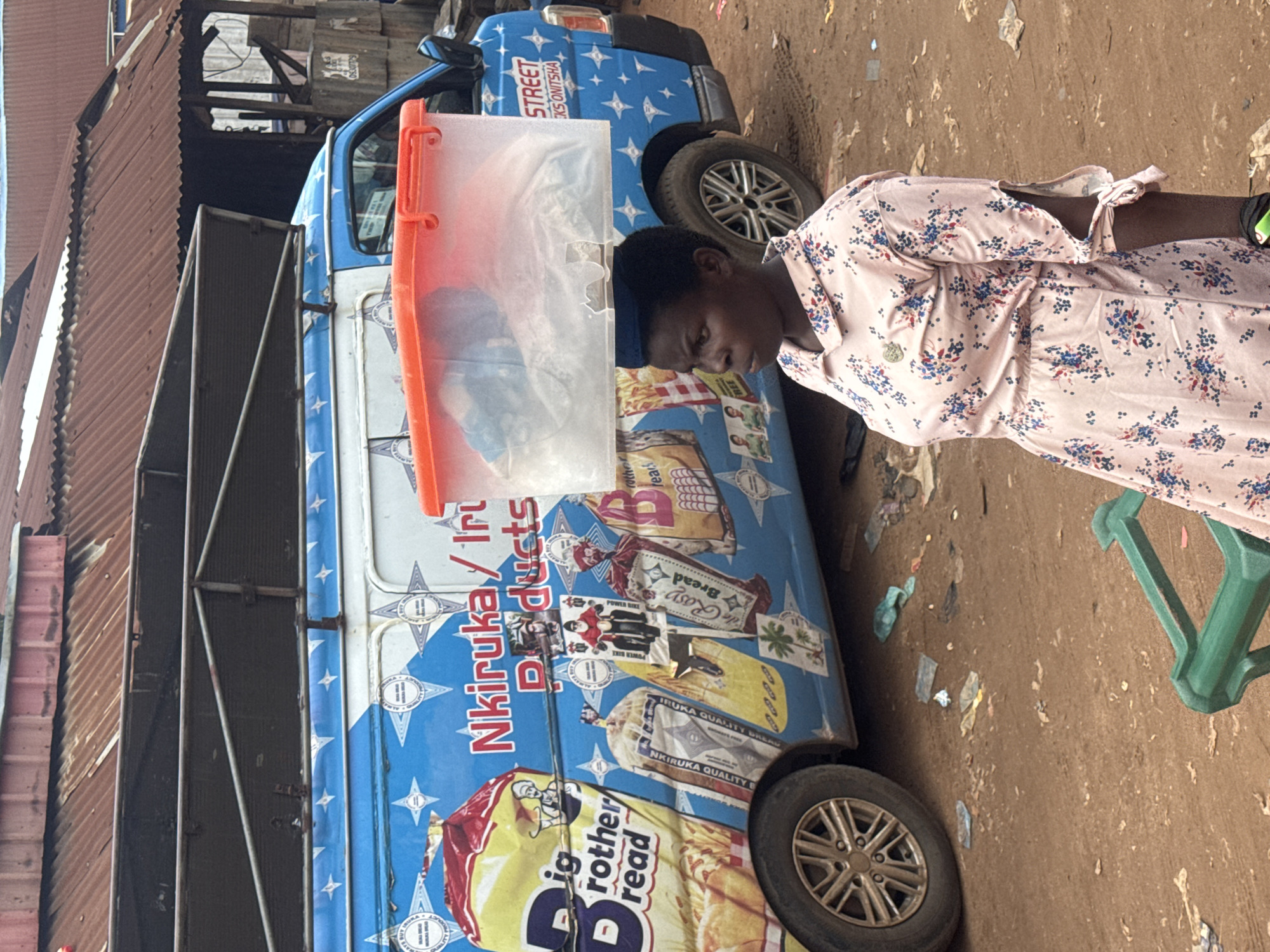 A woman with dark skin and short black hair carries a clear plastic container filled with white material on her head. She wears a light pink dress with a floral pattern of blue and red flowers and green leaves. Her gaze is directed to her right, with her mouth slightly open. She is standing in front of a blue van with white lettering and graphics, including images of bread products. The van's side displays the text "Nkiruka / Iri Products" and "STREET CKS ONITSHA". The environment appears to be a dusty, outdoor market setting with scattered debris on the ground. In the background, a corrugated metal roof structure is visible, suggesting a semi-built or informal commercial area. The overall scene depicts a moment of commerce and daily life in a Nigerian city, likely Nkpor.
