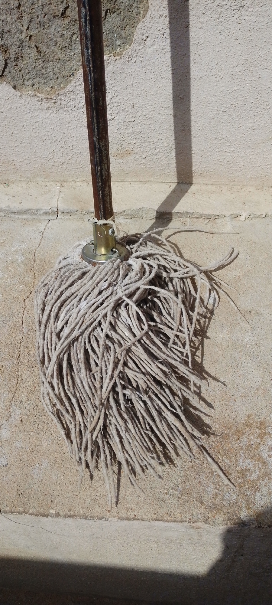 A well-used mop with a weathered metal handle leans against a textured, light-colored wall. The mop head is made of numerous frayed, off-white strands. A metallic connector piece attaches the mop head to the handle. The wall shows signs of age, with a rougher, greyish patch near the top and a horizontal crack running through it. The scene is illuminated by sunlight, casting a distinct shadow of the mop handle diagonally across the wall and ground. The ground in the foreground is a similar textured, light-colored surface. The image appears to be taken outdoors, possibly in an urban or outdoor setting.