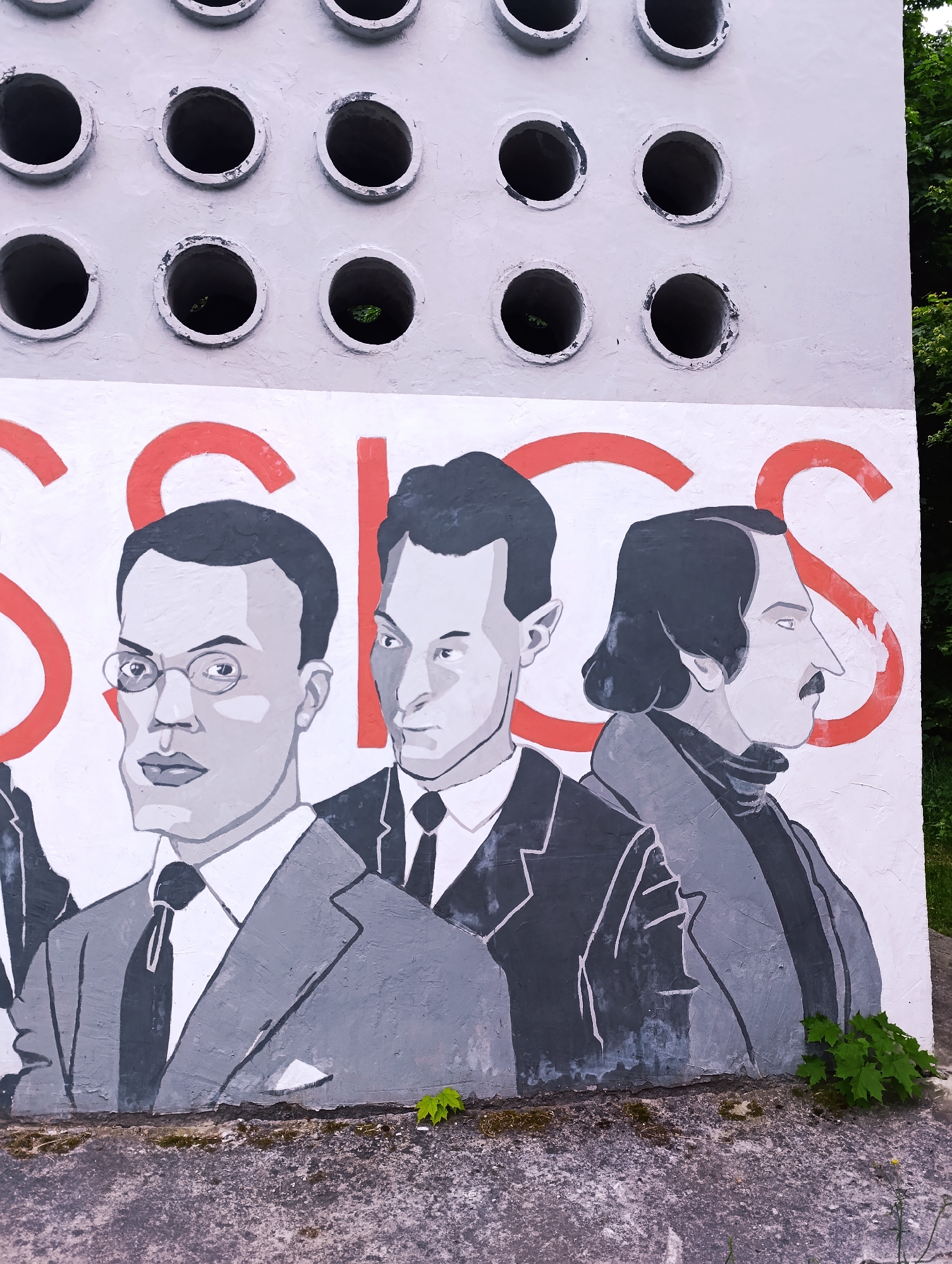 The image shows a mural in an unknown location in the city of Unknown, Unknown. The mural features three grey-scale portraits of men in suits, overlaid with red lettering that appears to spell "CLASSICS." The man on the left wears round glasses. Above the mural is a grey wall with multiple circular openings, some of which contain vegetation. The bottom of the mural rests on a concrete ledge with moss and small green plants growing on it. Green foliage is also visible on the right side of the image.