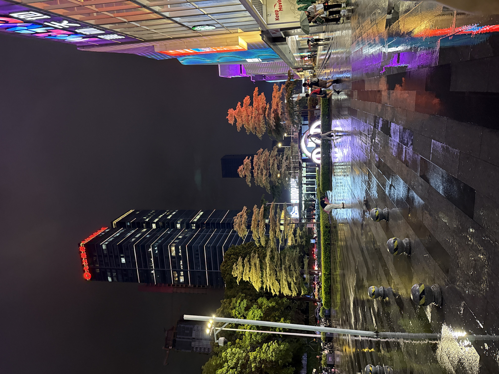 The image depicts a nighttime street scene in Tianhe District, China, following rainfall, evidenced by wet pavement reflecting the surrounding lights. A tall, modern skyscraper with red neon signage at its peak dominates the left side of the frame. In the foreground, several people are walking on the wet sidewalk, some holding umbrellas. The right side of the image features a brightly lit building with colorful neon advertisements, including what appears to be a Starbucks logo. Trees with distinctly tiered foliage are strategically placed along the street. The ground is paved with large, dark rectangular tiles, interspersed with lighter gray ones, creating a patterned walkway. Several bollard-like objects with yellow and black striped markings are positioned on the pavement. The sky is dark and cloudy, indicative of nighttime.