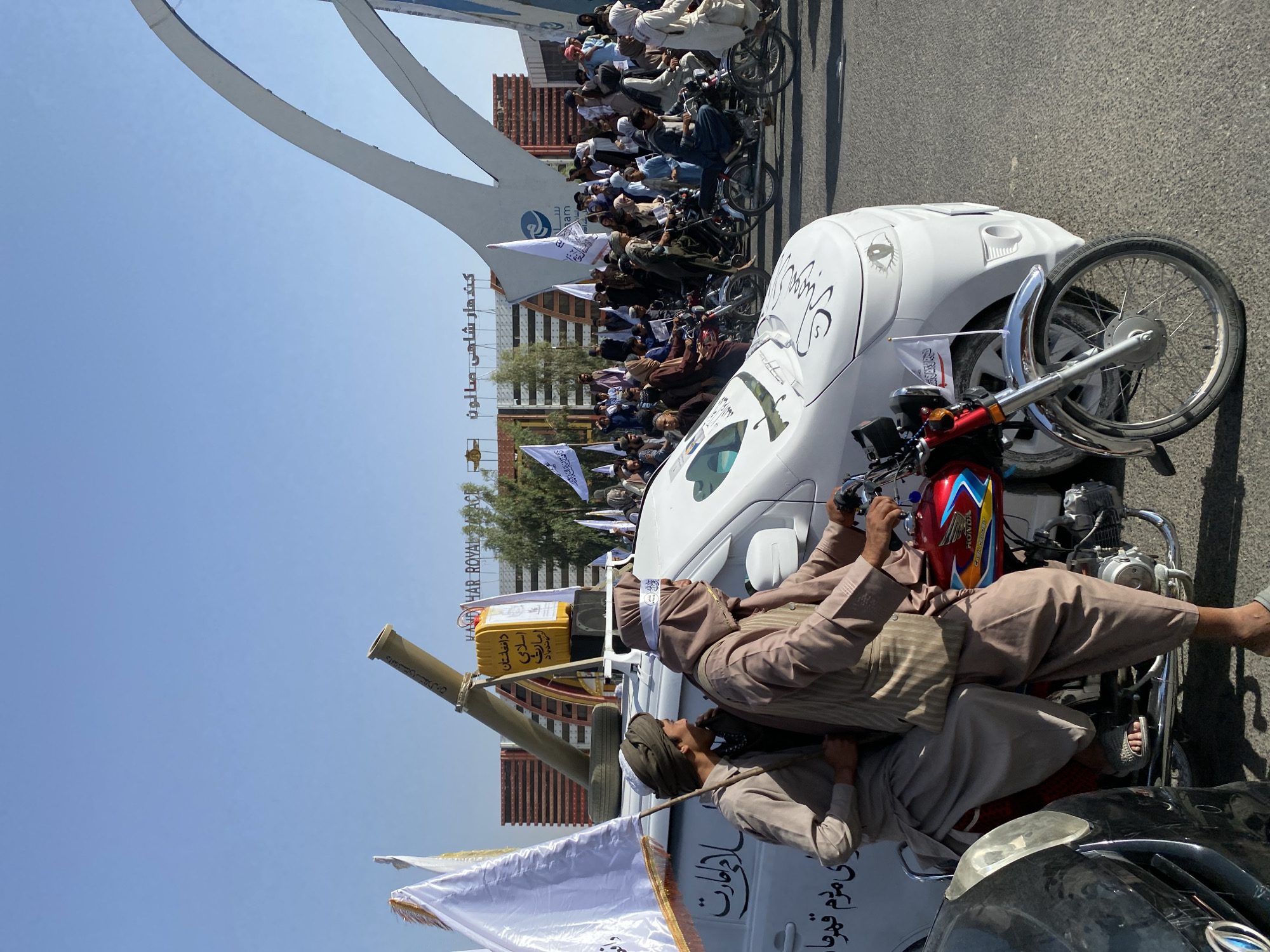 In the city of Kandahar, Afghanistan, a procession of people on motorcycles and in a white van moves along a paved road under a clear blue sky. The van, adorned with white flags and black Arabic script, carries a mortar on its roof. Two men, dressed in traditional Afghan clothing, sit on a red motorcycle in the foreground, one driving and the other holding a white flag. In the background, a large modern building with a white arched structure and signage, which reads "Kandahar Royal Palace" in English and Arabic, is visible. A crowd of people, many riding motorcycles and carrying flags, fills the street. The overall atmosphere appears to be that of a parade or demonstration.