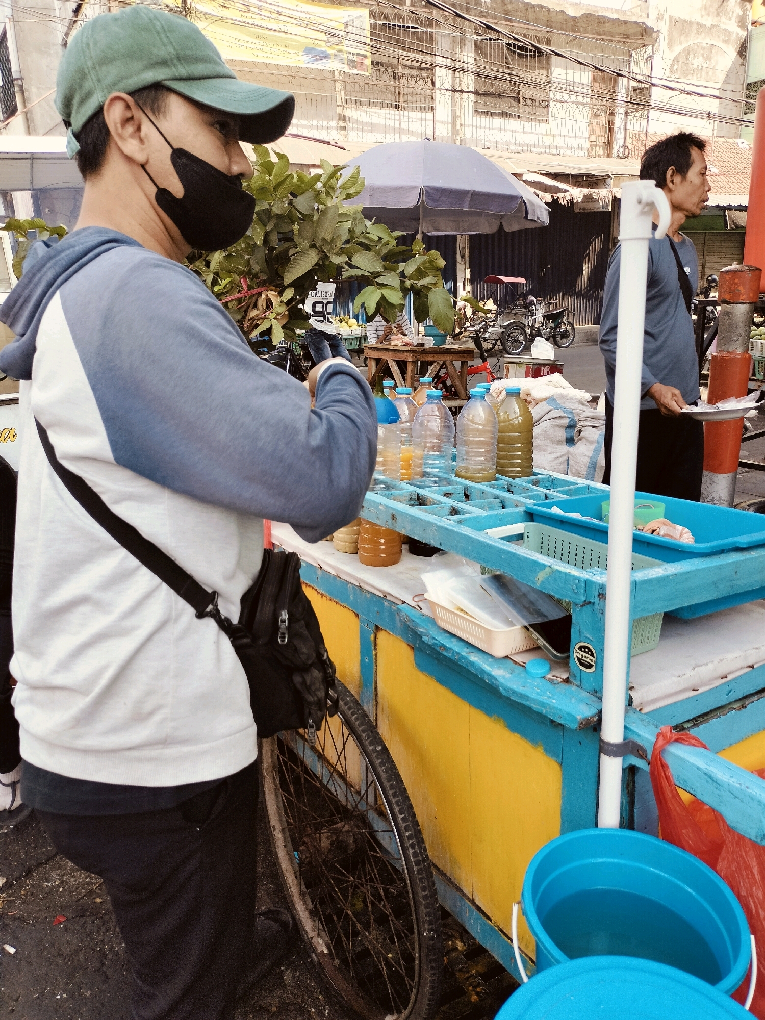 A man wearing a green cap and a black mask is standing by a food cart in Surabaya, Indonesia. He is wearing a blue and white hoodie and a black crossbody bag. The food cart is painted blue and yellow and has a wheel on the side. There are several bottles of drinks on the cart, as well as some containers. In the background, another man is standing and holding a plate. The scene appears to be a street market or a roadside stall.