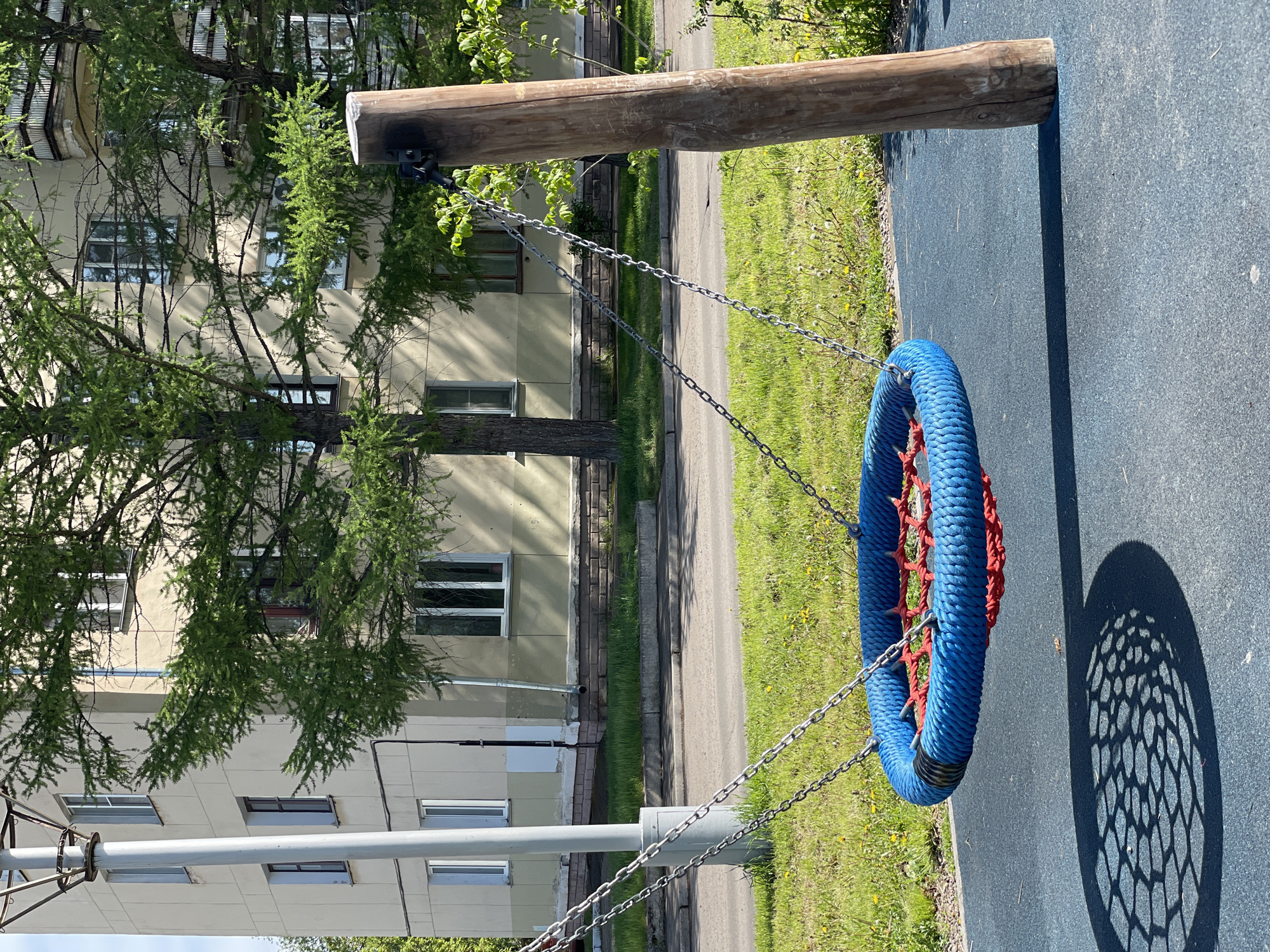 The image depicts a blue and red circular swing hanging from a wooden beam and chains in Zheleznogorsk, Russia. The swing's shadow falls on a blue paved surface. A light-colored building with numerous windows is visible in the background, partially obscured by a tree with dense green foliage. A section of asphalt road and grassy area are also present.