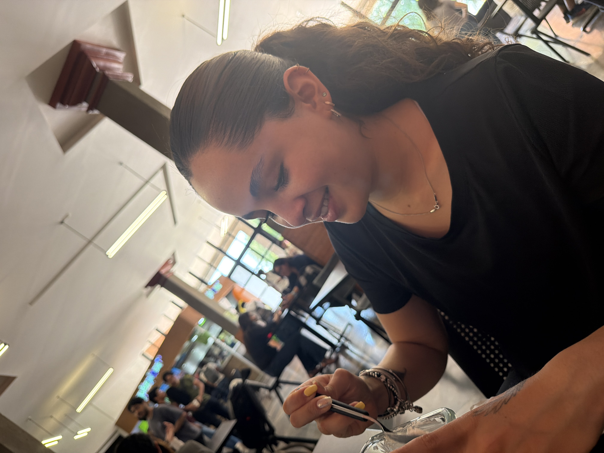 An eye-level shot, slightly tilted, captures a young woman smiling and looking down while holding a utensil over a clear glass container. She is positioned in the foreground, with her right side facing the viewer. Her dark hair is pulled back in a high ponytail, revealing multiple small earrings in her right ear and a simple silver necklace. She wears a black t-shirt and has several bracelets on her left wrist, with her fingernails painted yellow and silver. A tattoo is subtly visible on her left forearm.
The setting appears to be a casual indoor space, possibly a cafeteria or restaurant in San Andrés Cholula, Mexico, given the architectural style. The background features several blurred figures of people seated at tables, suggesting a busy environment. Large windows allow natural light to illuminate the space, complemented by long, rectangular fluorescent lights fixed to the ceiling. The overall atmosphere is relaxed, with the woman's gentle smile conveying a sense of contentment. No discernible text is visible in the image.