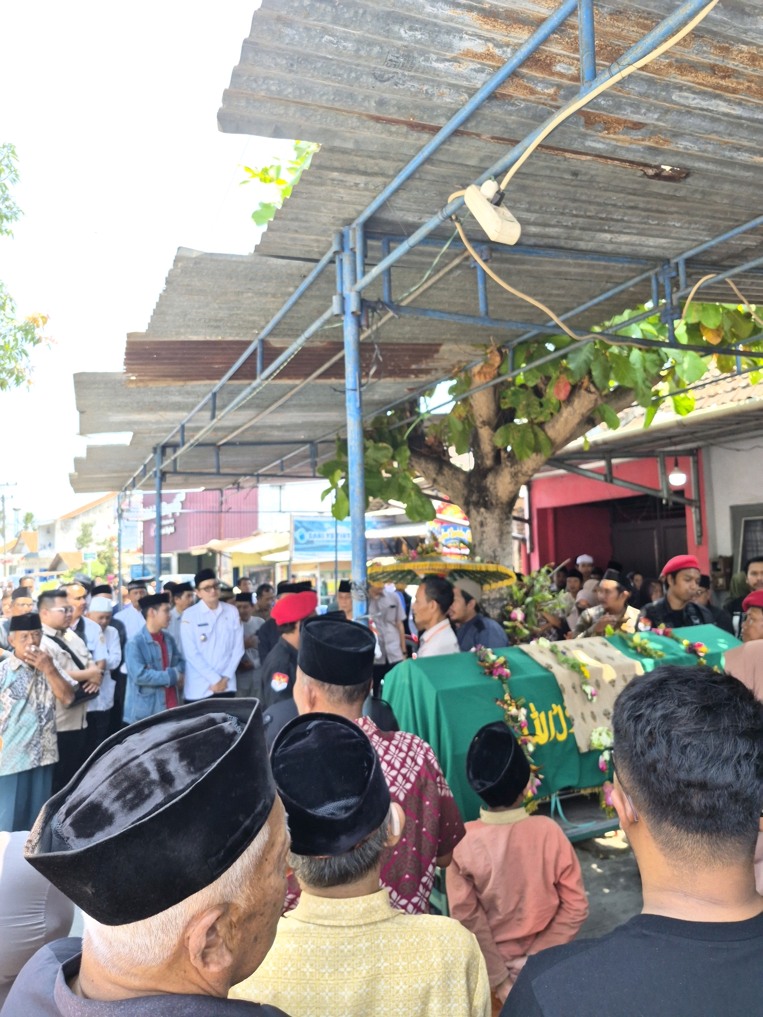 The image depicts a funeral procession in Semawung Daleman, Indonesia. A green-covered casket, adorned with flowers and Arabic calligraphy, is positioned under a corrugated metal roof supported by blue metal poles. Several men, many wearing traditional Islamic caps (peci) and some with red berets, are gathered around the casket. Some individuals are holding umbrellas. In the background, buildings, trees, and a crowd of onlookers are visible. The scene suggests a solemn and formal occasion.