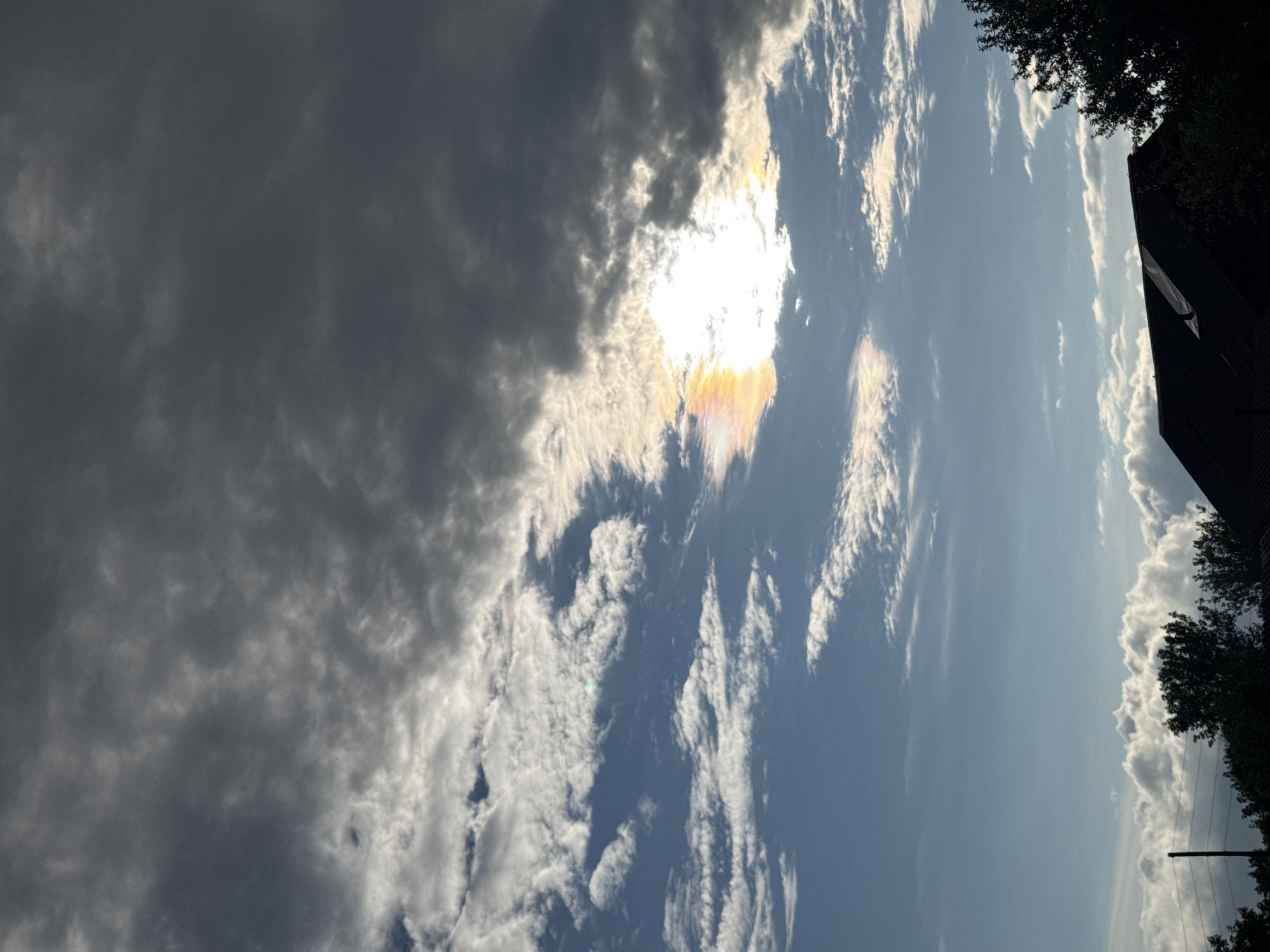 The media file shows a sky with dramatic clouds and the sun. The sun is partially obscured by dark, heavy clouds on the left side of the frame, with rays of light breaking through. There are also thinner, wispy clouds in the sky, some of which are illuminated by the sun, creating a rainbow-like effect. The sky transitions from dark and stormy on the left to a bright, clear blue on the right. In the upper right corner, a silhouette of trees and a dark building is visible, suggesting an outdoor setting. The orientation of the image is portrait. The weather appears to be changing, possibly transitioning from a storm or heavy cloud cover to clearer conditions. The scene evokes a sense of awe and the power of nature. There is no visible text in the image.