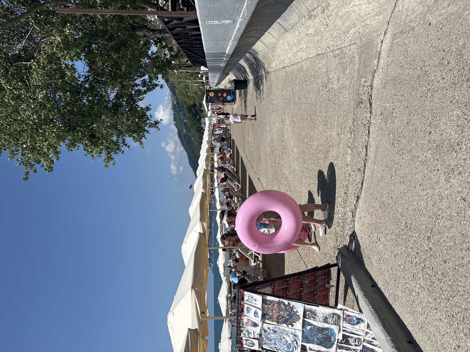 The image depicts a sunny day on a beach promenade in Sochi, Russia. White beach umbrellas are lined up along the shore, providing shade for beach chairs and people. In the foreground, two young individuals are seen standing on a concrete path, one holding a large pink inflatable ring. To the left, a display rack features various artwork or temporary tattoos. Further down the path, other beachgoers are visible, some walking and others relaxing. A child in a light-colored shirt walks away from the camera. A trash can and a vending machine are also present on the right side of the promenade, which runs alongside a decorative concrete barrier. In the background, a lush green hillside with buildings suggests a coastal resort town. The sky is clear and blue.