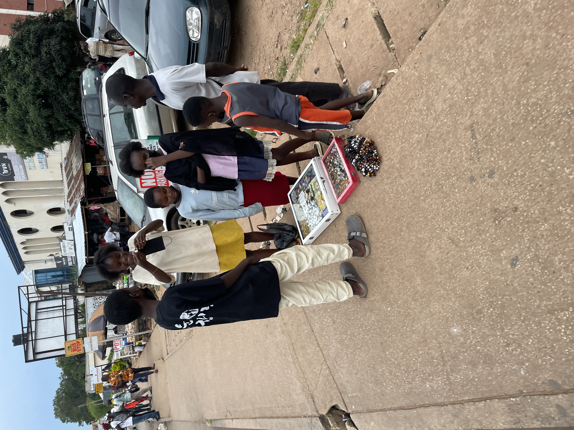 The image captures a street scene in Jos, Nigeria, likely near a bus stop given the presence of a visible "BUS STOP" sign and a kiosk with "POSTAL SERVICE" signage in the background. The foreground is dominated by a group of individuals, primarily children and young adults, gathered on a concrete sidewalk. They appear to be in the process of displaying or examining items for sale.
In the center of the group, a young person in a black t-shirt with white graphics and light-colored pants stands with their back to the camera. To their right, a young person in a cream-colored t-shirt and yellow skirt is holding a black face mask and looking towards the person in the black t-shirt, seemingly engaged in a conversation or transaction. Another young person, dressed in a light blue long-sleeved shirt and red skirt, stands to the left of the person in the black t-shirt, also looking at the items displayed. A young person in a dark outfit with a pink undershirt stands between the two central figures, also looking down. To the right of this group, an adult male in a white t-shirt and dark pants stands observing. Further to the right, a young person in an orange and gray sleeveless shirt and orange-striped shorts is visible.
On the ground in front of the group are several items laid out for sale. These include a white display case filled with various accessories, possibly jewelry or trinkets, and a red tray containing similar items. A pile of beaded bracelets, in dark and lighter colors, is also present.
The environment shows a typical urban setting with buildings in the background, including one with a large, blank billboard structure. Several vehicles are parked or driving on the street adjacent to the sidewalk, including a silver station wagon and a dark green sedan. The ground is a mixture of concrete and dirt, with some visible cracks in the sidewalk. The sky appears overcast, suggesting a cloudy day. The overall scene depicts a lively street commerce environment.