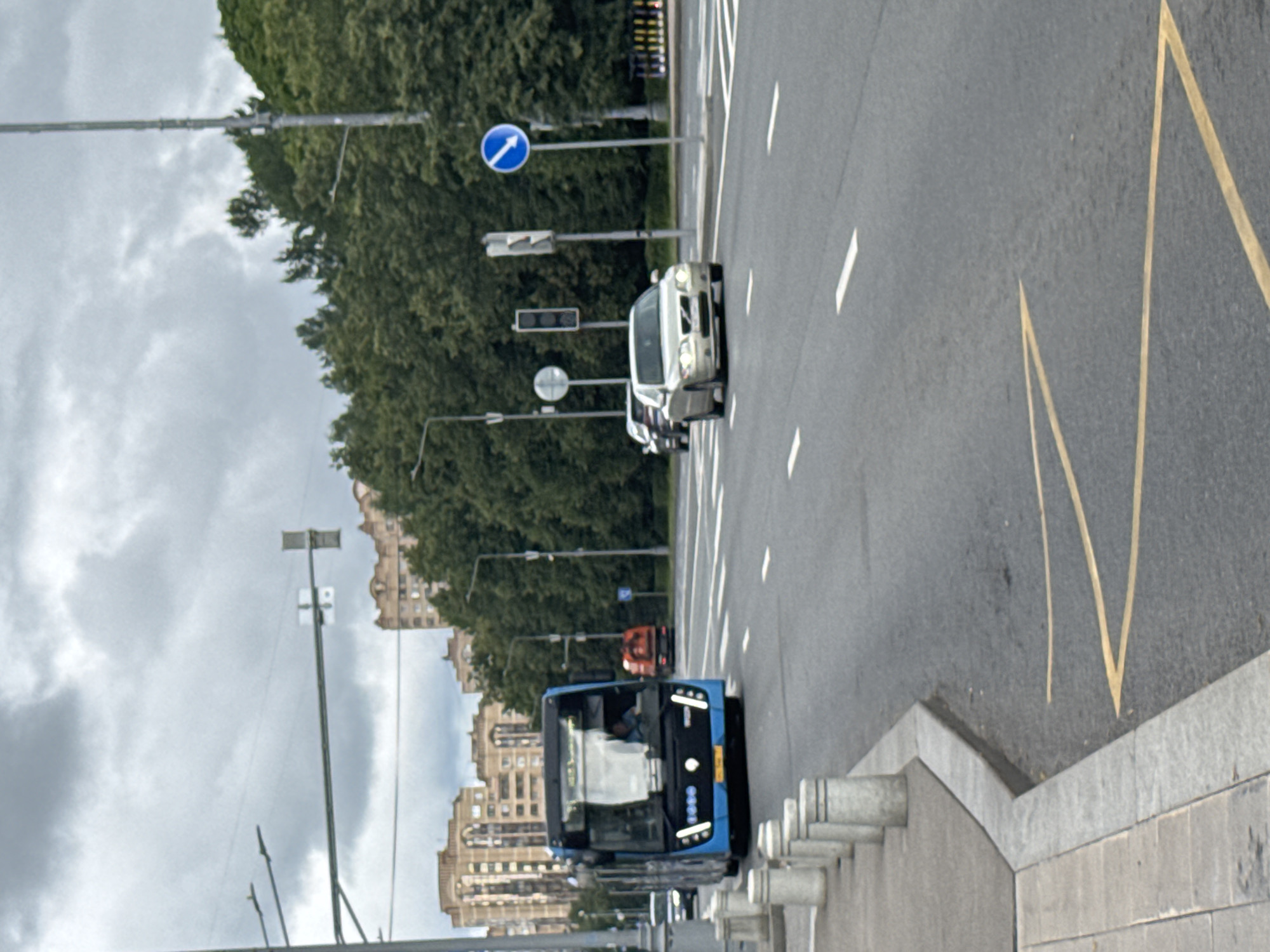The image captures a view of a street in Zelenograd, Russia, under a cloudy sky. A blue bus with illuminated headlights is in the foreground, driving on the right side of the road. To the right of the bus, a silver sedan is also in motion. In the background, several buildings, mostly residential with light-colored facades, are visible behind a dense line of green trees. Traffic signals and streetlights are present, along with road markings, including white lane dividers and yellow directional arrows on the asphalt. A sidewalk with bollards lines the left side of the road.