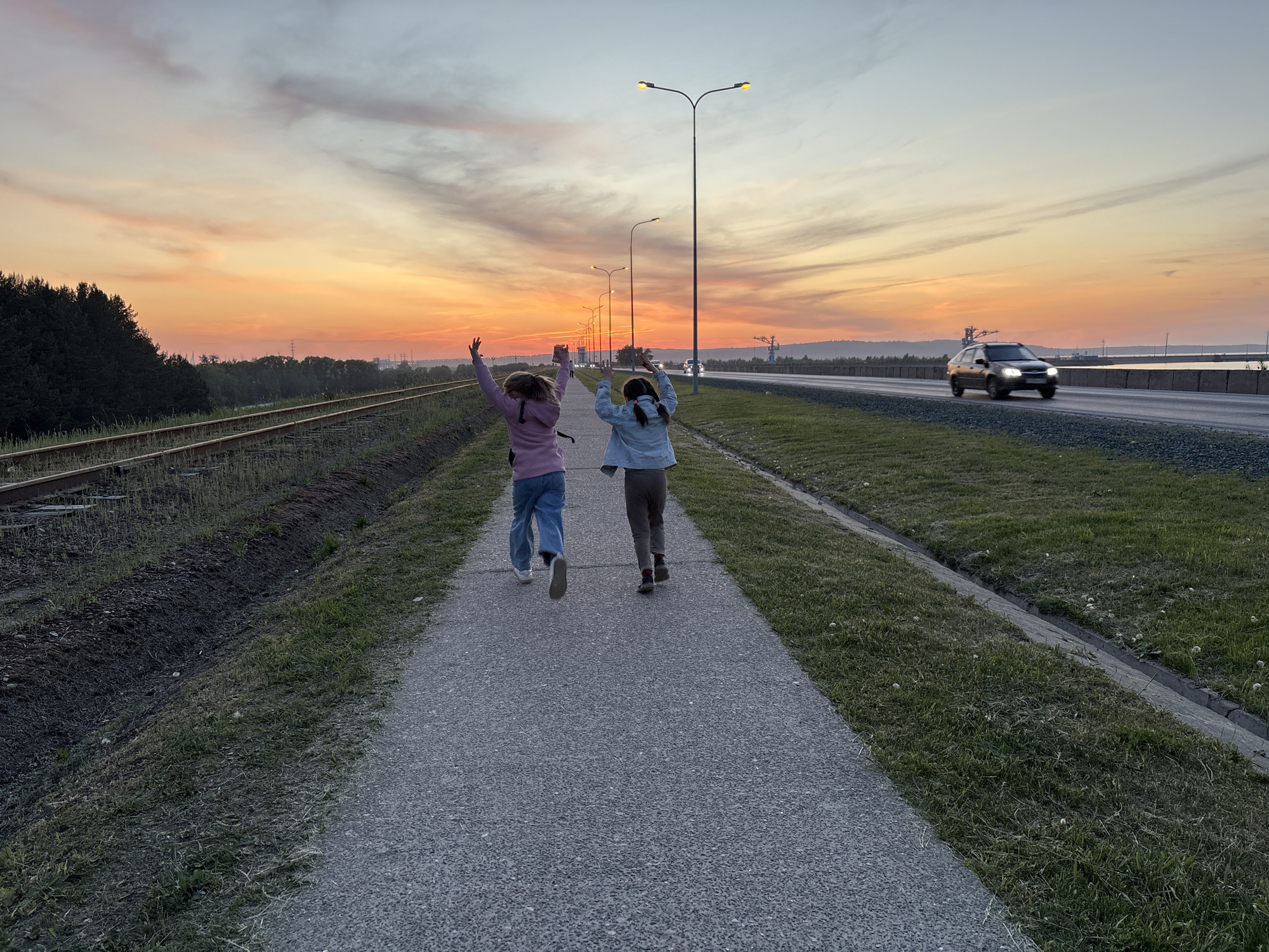 Two young girls are running down a grey paved path in the city of Chaikovsky, Russia. The girls are facing away from the camera with their arms raised in the air. The girl on the left is wearing a pink jacket and blue jeans, while the girl on the right is wearing a light blue jacket and brown pants. The path runs parallel to a set of railroad tracks on the left, and a road with a car traveling on it on the right. There are street lights lining the road. The sky is orange, indicating sunset.