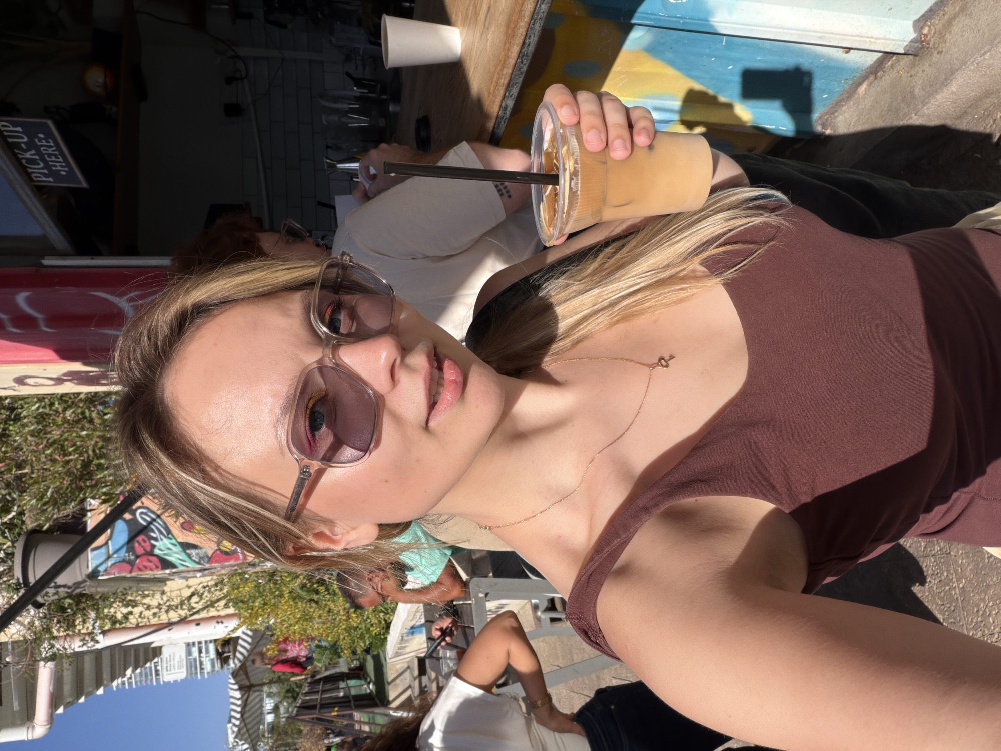 A young woman with blonde hair and sunglasses holds a coffee drink in a plastic cup with a straw. She is wearing a brown tank top and a gold necklace. Other people are visible in the background. The location is an outdoor cafe in Tel-Aviv, Israel, indicated by the surroundings.