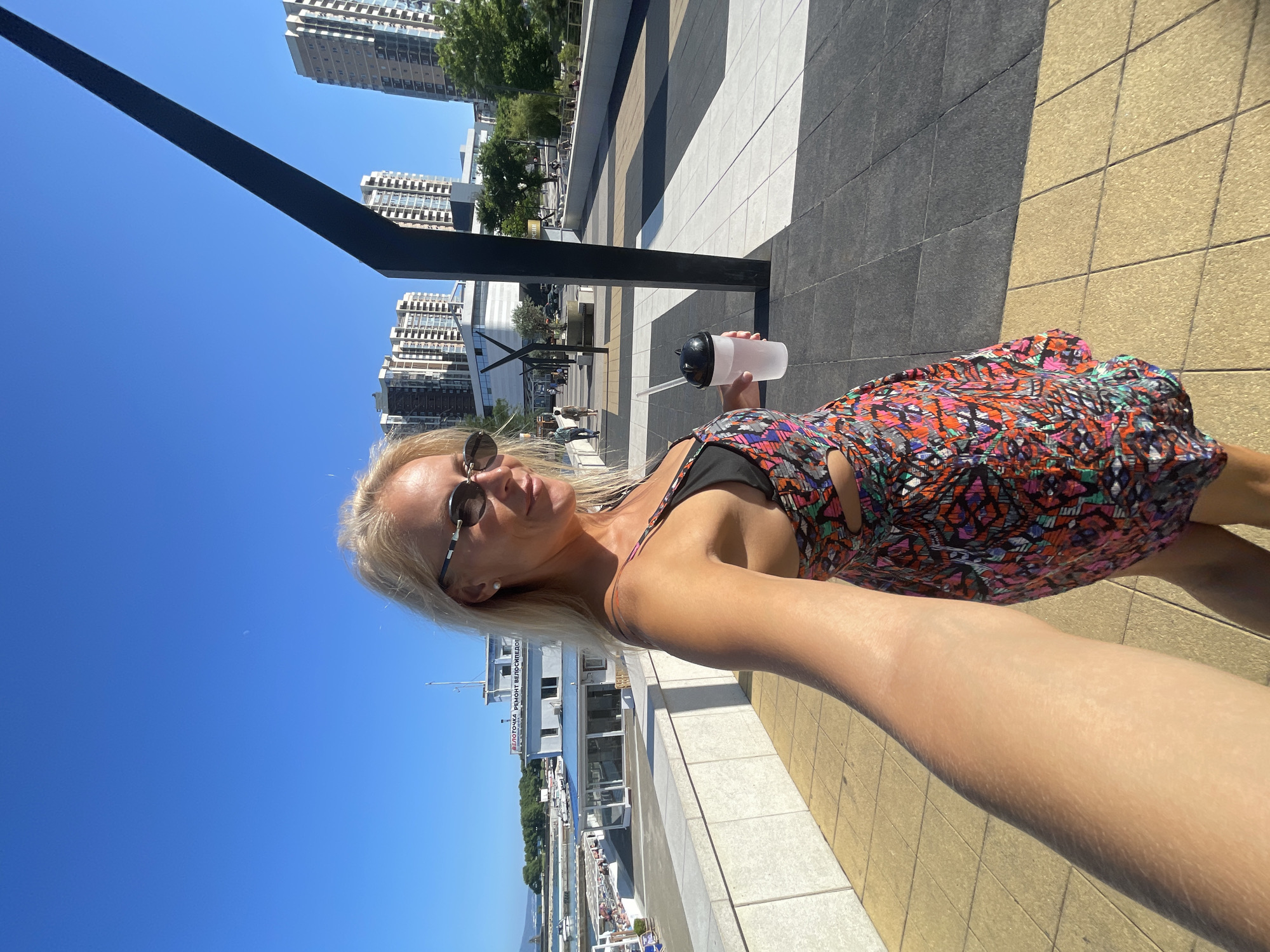 A woman is taking a selfie outdoors on a sunny day. She has blonde hair, is wearing sunglasses and a colorful patterned dress. She is holding a clear tumbler with a black lid and a straw. The setting appears to be a modern urban waterfront area with tall buildings and a clear blue sky. The ground is paved with large square tiles in alternating dark grey and beige. A dark metal pole is visible in the foreground. In the background, there are more modern buildings, trees, and a body of water with boats. One building has Russian text that reads "ВЕЛОСИПЕДНЫЙ РЕМОНТ" which translates to "BICYCLE REPAIR". The overall mood is relaxed and summery.