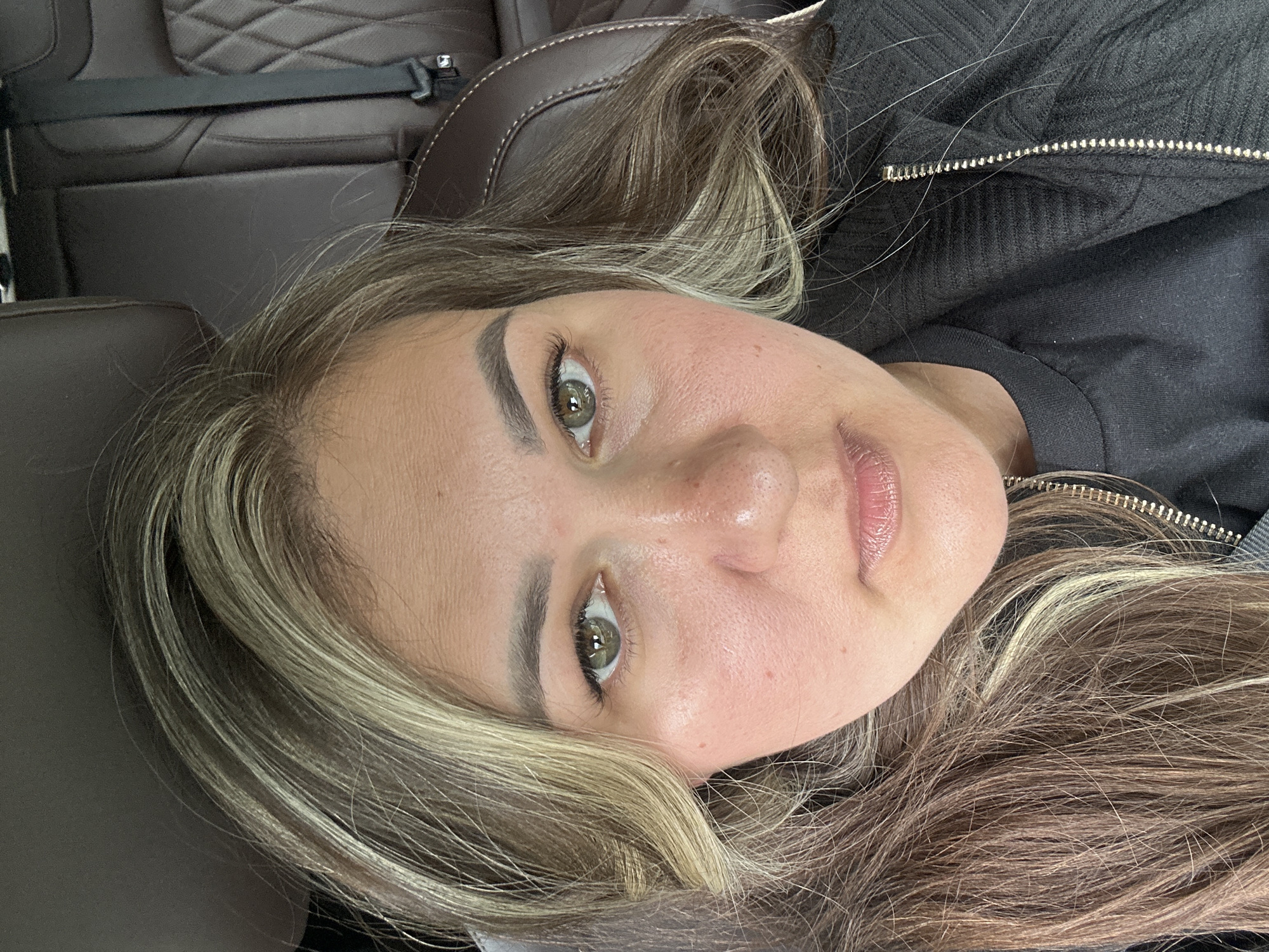 A close-up, angled portrait shows a woman with fair skin and light brown hair with blonde highlights. She has light green eyes, defined eyebrows, and slightly parted lips. She wears a dark-colored jacket with a visible zipper. The background includes a brown leather seat with quilted stitching and a seatbelt. The image doesn't give enough information to place the subject in Новая Казмаска, Russia.