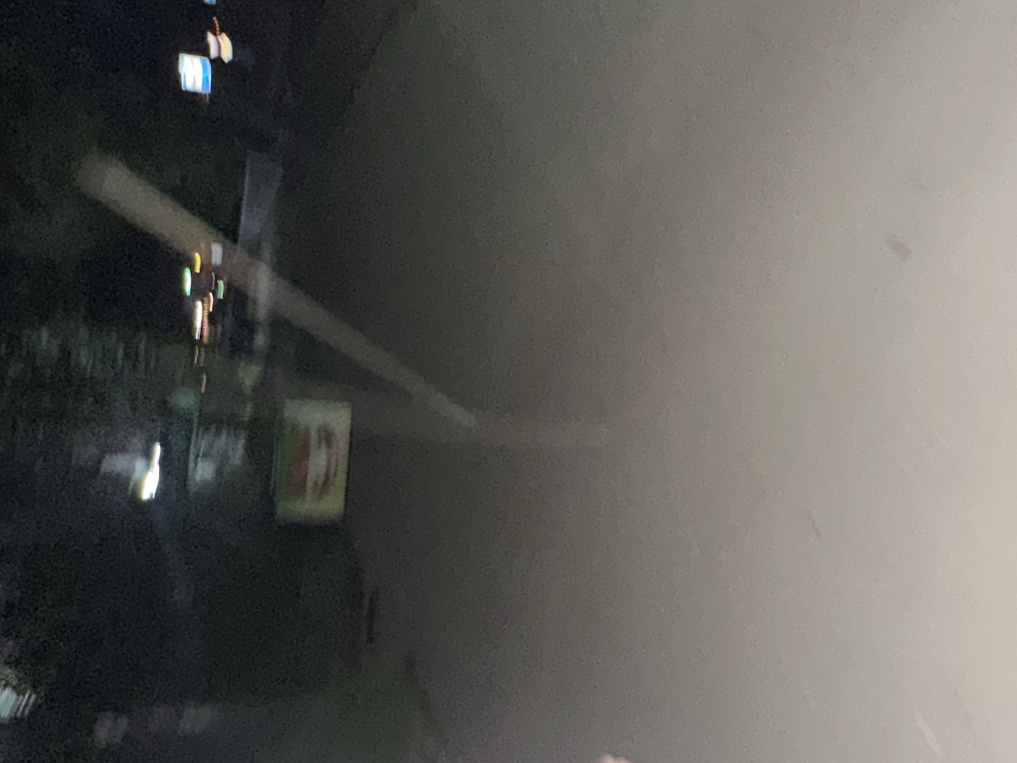 The image appears to be a night-time photograph taken through a window, possibly from inside a vehicle. The overall scene is dark with blurred lights and reflections.
In the background, on the left side, there are several illuminated objects that suggest an urban or roadside setting. These include what look like streetlights or building lights, with some exhibiting blue and white hues. Below these, a rectangular sign with red and white elements is visible, possibly an advertisement or a marker. There are also streaks of light, indicating motion blur, suggesting the photo was taken while moving.
The majority of the image is filled with a soft, out-of-focus glow, likely a reflection on the glass surface. This reflection is lighter and more diffuse than the scene behind it.
No clear subjects like people or specific objects are distinctly identifiable due to the blur and low light conditions. The setting appears to be outdoors at night, in an area with artificial lighting. The weather is not discernible.
There is no visible text that can be clearly read. The overall mood is somewhat abstract and atmospheric due to the lighting and blur. The location context of Birobidzhan, Russia, is noted but not visually evident within the image itself.