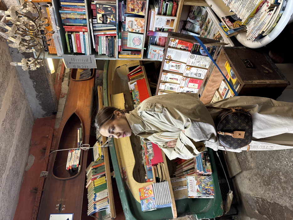 A female individual stands in the interior of Libreria Acqua Alta, a bookstore located in Venice, Italy. She is positioned slightly right of center, facing left with a neutral expression, and her arms are crossed over her chest. She wears a light beige trench coat and carries a brown, circular Louis Vuitton monogrammed cross-body bag with a tan leather tag on her right hip.
The environment is a cluttered bookstore characterized by unconventional shelving. Two wooden boats, appearing to be canoes, are integrated into the wall as bookshelves, filled with stacked books. One canoe is natural wood, the other painted green. Additional books are displayed on standard wooden shelves, in an open wooden chest with a slanted display top, and within a white bathtub visible in the lower right foreground. A multi-armed, ornate chandelier with clear glass components hangs from the exposed, concrete ceiling in the upper right. The walls are partially painted red with visible wear and exposed plaster. The floor is an uneven concrete or stone surface.