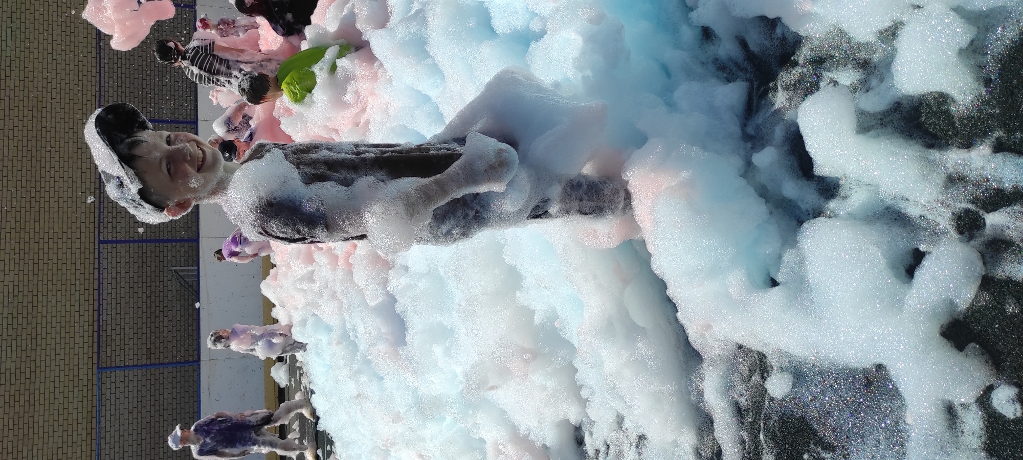 The image shows a group of children playing in a large amount of white and pink foam. The children are dressed in casual clothing. One boy in the foreground is wearing a dark t-shirt and a baseball cap, and he is smiling at the camera. Other children are visible in the background, some partially obscured by the foam. The foam appears to be a recreational activity, possibly a foam party, set up outdoors. The background suggests an urban or semi-urban environment, with a brick building and a blue fence visible. The ground beneath the foam is dark and appears to be asphalt. The image was taken on a sunny day, as indicated by the bright lighting and shadows. The location is identified as Noviy, Russia.