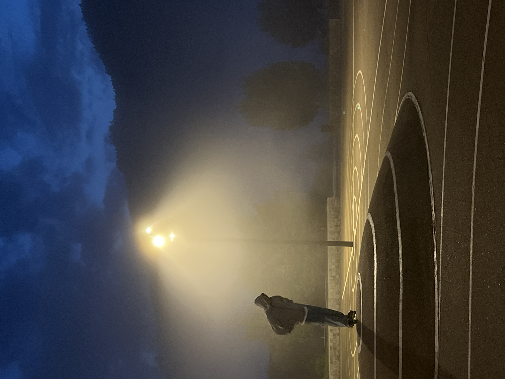 This is a vertical, low-angle, eye-level shot taken at night in Divnogorsk, Russia. A person wearing a hooded jacket and jeans stands on a paved surface with white lines, possibly a basketball court or running track, marked with concentric arcs. The person is looking towards a bright light source, which appears to be a streetlamp illuminating a dense fog. The fog creates a dramatic, ethereal effect, obscuring the background and casting a warm glow around the light. To the left of the frame, a dark, mountainous landscape is visible, silhouetted against a deep blue, cloud-filled sky. The overall mood is solitary and mysterious, with the fog and dim lighting contributing to a sense of isolation and contemplation. There is no visible text in the image.