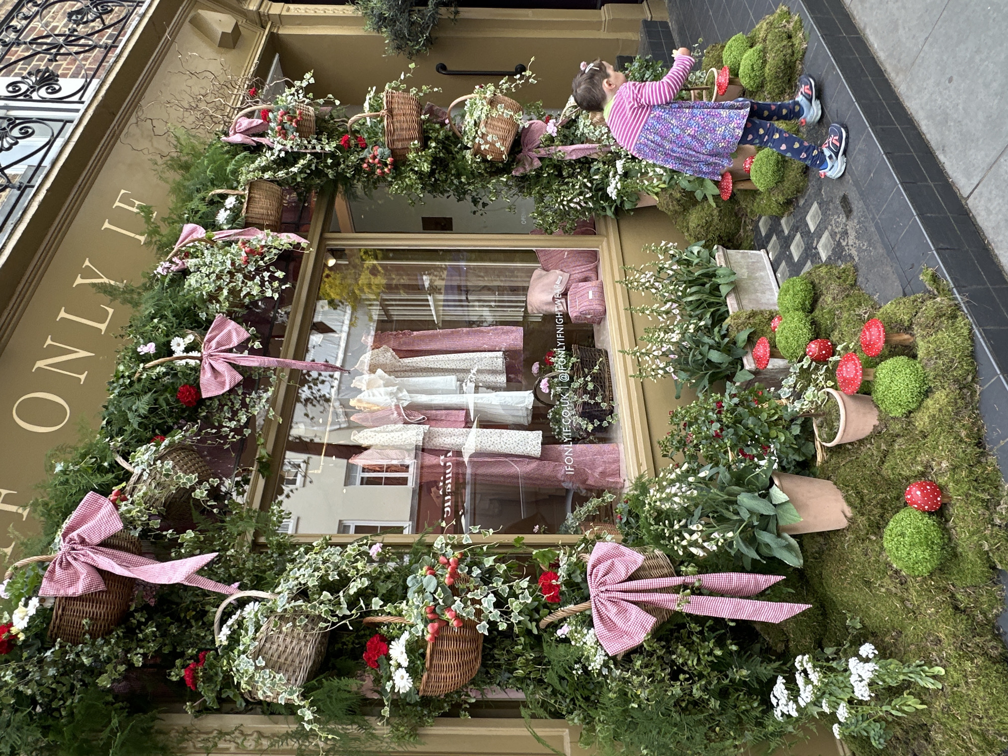 A shopfront display in London, United Kingdom, featuring an abundance of greenery, flowers, and wicker baskets adorned with red and white checkered bows. The display is centered around a window showcasing fabric and garments. A young child stands on the right side, interacting with the display. The sidewalk is visible at the bottom. The building's architecture is visible.