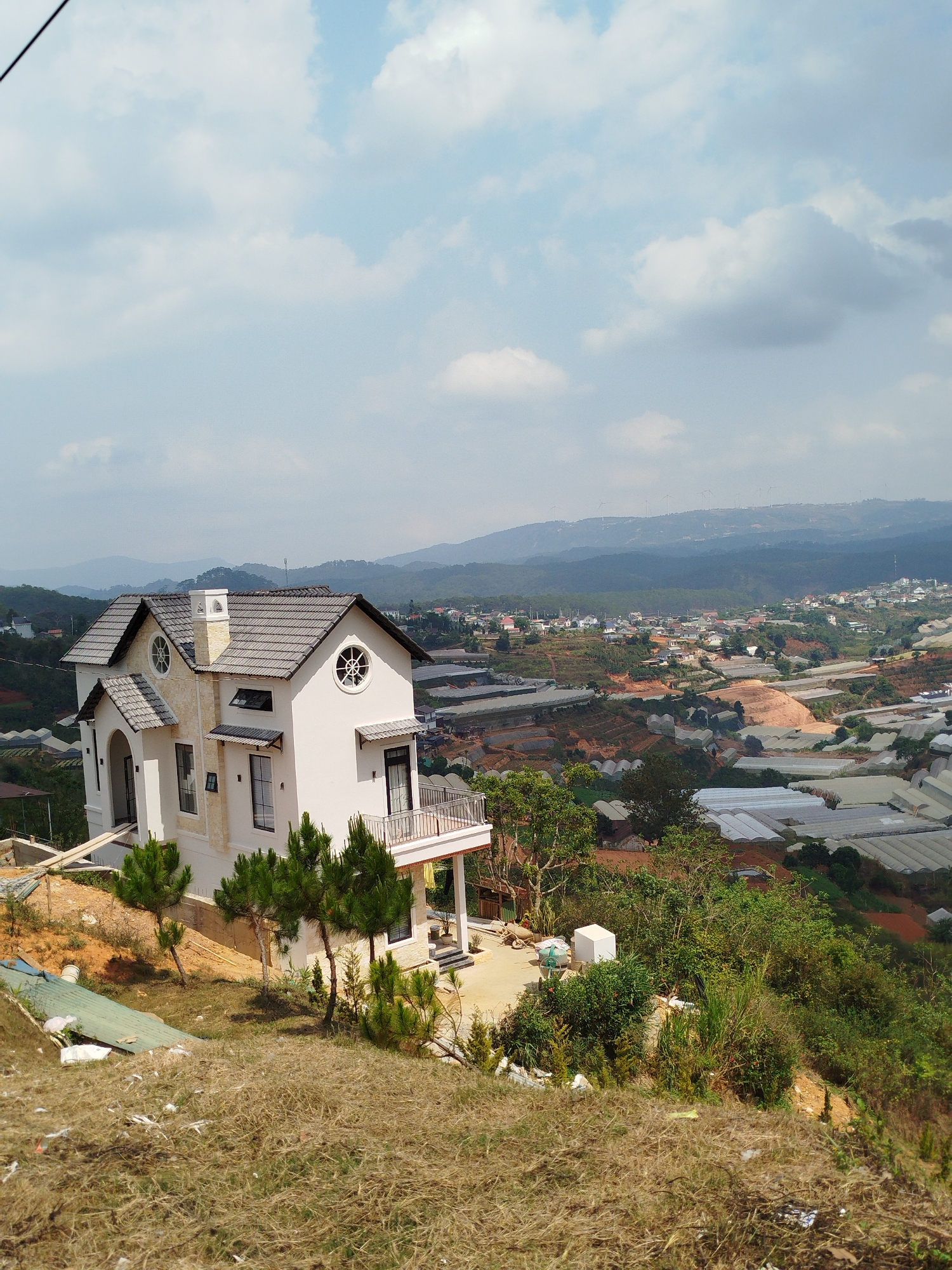 The image shows a two-story white house with a gray tiled roof situated on a hillside in Đa Thọ, Vietnam. The house features a balcony, arched doorways, and circular windows. The surrounding landscape is a mix of dry grassy slopes, evergreen trees, and other vegetation. In the background, there are cultivated fields, greenhouses, and other buildings, indicating a populated area. The sky is partly cloudy.