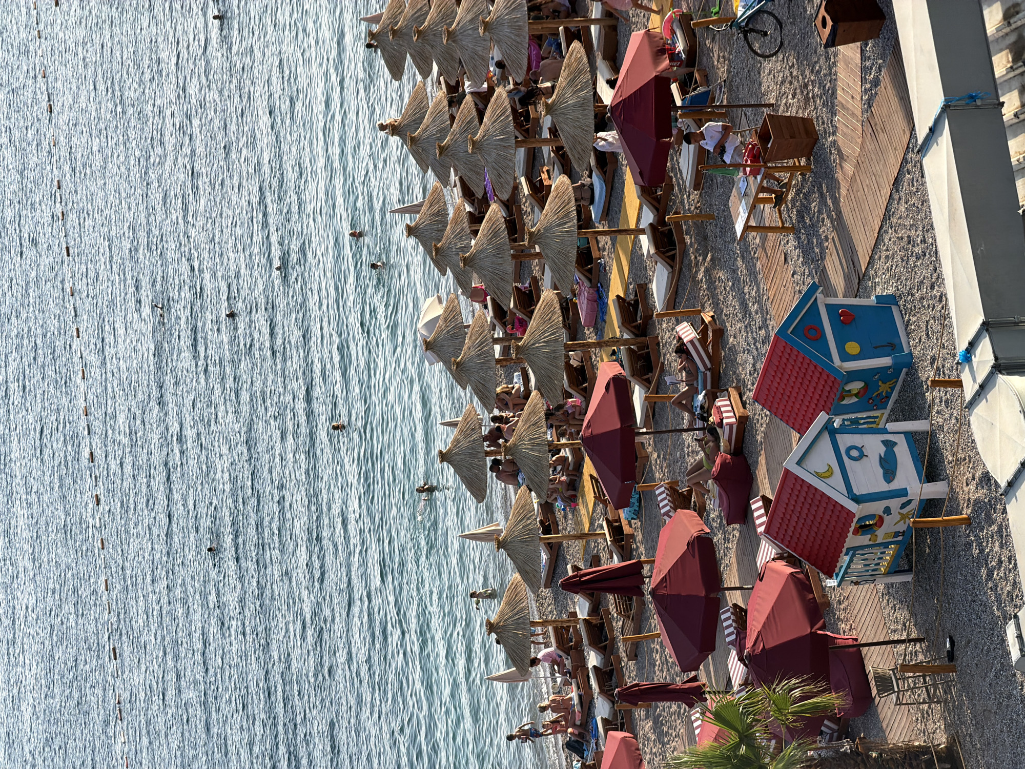 The image depicts a sunny beach scene in Boreti, Montenegro, with the Adriatic Sea in the background. The water is calm, with several individuals swimming and enjoying the sea. The beach itself is filled with lounge chairs and thatched umbrellas, providing shade for beachgoers. Some people are seated on lounge chairs, while others are walking or relaxing on the sand. In the foreground, there are two colorful, playhouse-like structures, likely for children. A person is seated at a small wooden table, possibly attending to a booth. A bicycle is parked nearby. The overall atmosphere is one of leisure and relaxation on a warm summer day.