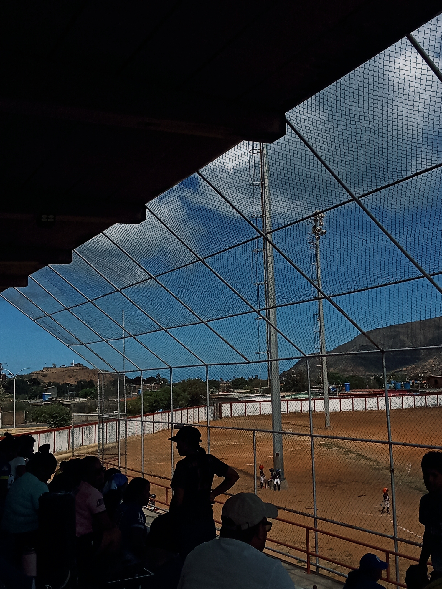 The image shows a baseball game in progress, viewed from the stands of a baseball field. People are watching the game from the stands. A chain-link fence surrounds the field. Behind the outfield fence, there is a low mountain and some buildings. The location is Juan Griego, Venezuela, based on the visual evidence of typical Venezuelan coastal scenery and baseball field architecture.