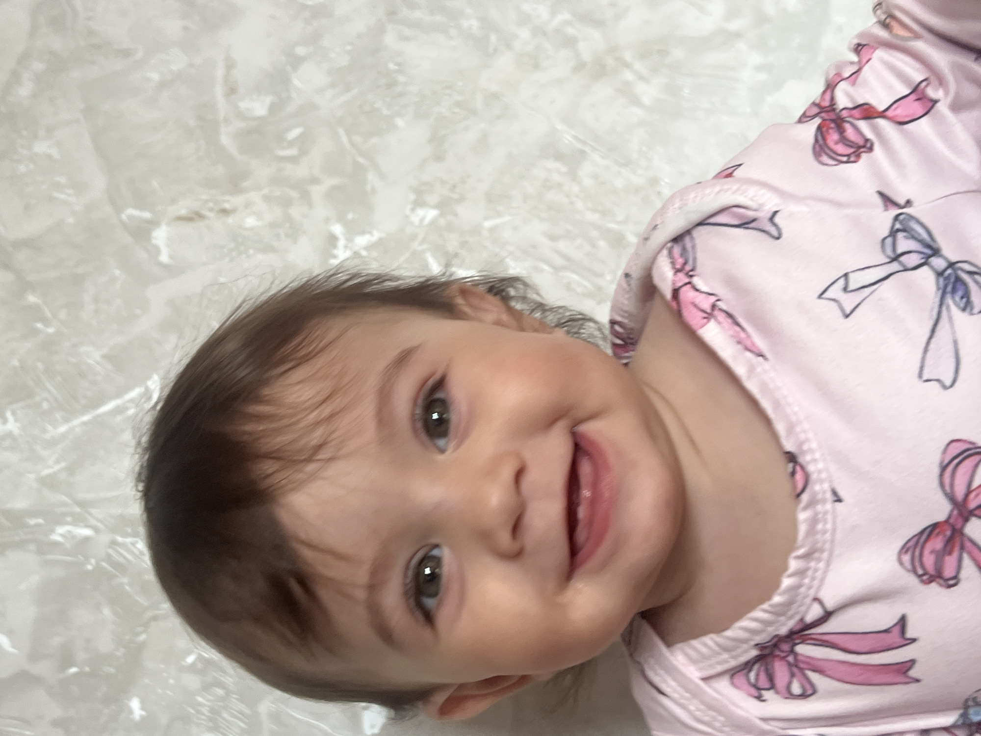 A close-up, slightly angled shot shows a baby with dark brown hair, hazel eyes, and fair skin, smiling with their mouth open, revealing their gums. The baby is wearing a light pink garment with bow patterns. The background is a soft, light-colored texture that could be a blanket or similar surface. The image does not show any indication of the city of Elena, Bulgaria.