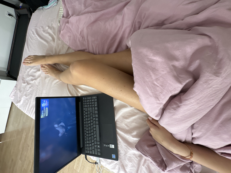 The image captures an overhead view of a person relaxing in bed, engaged with a laptop. The main subject is a person whose legs and one arm are visible. The person's bare legs, from mid-thigh down to the feet, are resting on light-colored bed sheets, possibly light pink or off-white. The visible arm, from the elbow down, rests gently on a light pink duvet or blanket that covers the rest of the person's body. A delicate, possibly braided or beaded bracelet is worn on the visible wrist.
A black laptop is placed on the bed to the left of the person's legs, open and turned on. White charging cables are visible near the laptop. The laptop screen displays a video or stream with a prominent blue banner at the top. The banner contains text in Cyrillic script: "ВСЕГО" (VSEGO, meaning "TOTAL") and "399 Р/МЕС" (R/MES, meaning "399 Rubles per month"). Below this, partially obscured, is the word "скидка" (skidka, meaning "discount"). The rest of the screen shows a dark scene, possibly from a movie or TV show, featuring a figure that appears to be a person, possibly in a white uniform or shirt, against a darker background.
The bed itself is covered with light sheets and a soft, crumpled pink duvet. To the left of the bed, a dark-colored treadmill is partially visible, indicating an indoor setting. Next to the treadmill, on a light wooden floor, there is a white piece of furniture, possibly a dresser or nightstand. The overall ambiance suggests a comfortable and relaxed personal space, likely a bedroom, with the person enjoying leisure time. The lighting in the image appears soft and natural, consistent with an indoor setting during the daytime or early evening.
