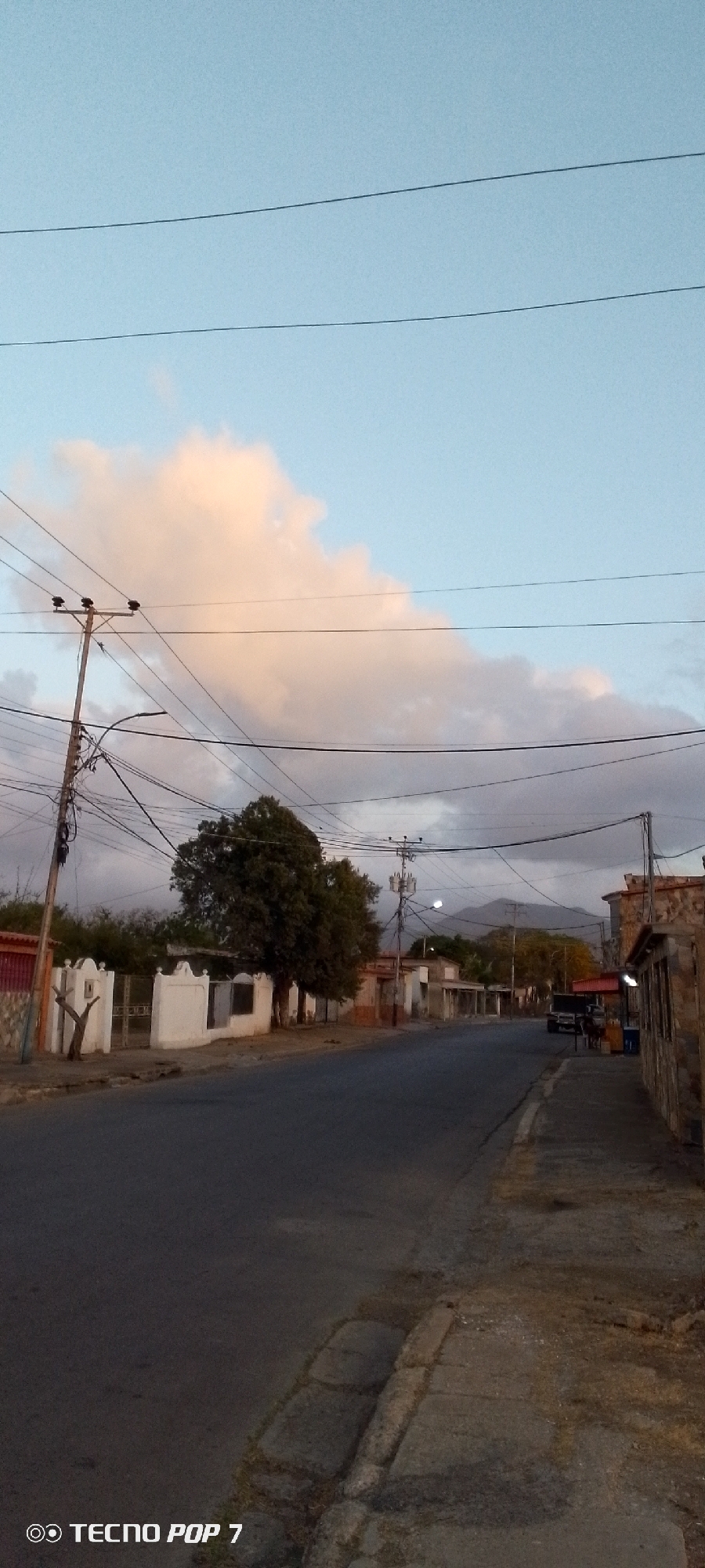 A street in Altagracia, Venezuela. The asphalt road stretches between buildings and houses on either side. Utility poles with numerous wires run along the roadside. A large tree stands near a white wall. In the background, mountains are visible under a partly cloudy sky. A vehicle is parked further down the street.