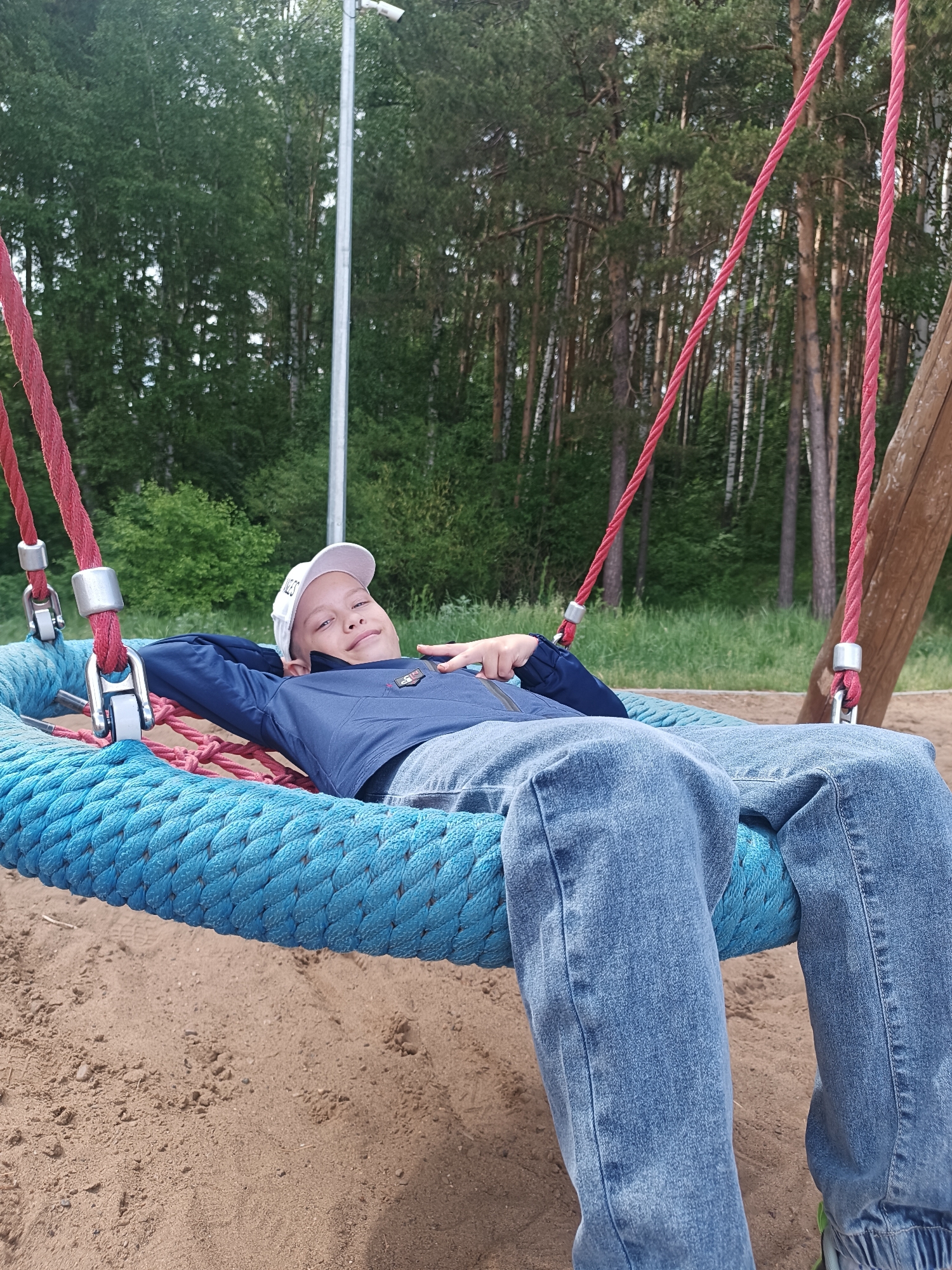 A young boy in a white baseball cap, navy jacket, and jeans lies back on a blue rope nest swing suspended by red ropes with metal hardware at a playground in Chaikovsky, Russia. The swing is situated on a sand surface. Behind the swing, a variety of trees form a backdrop, including pines and birches. A metal pole is visible on the left edge of the frame. The boy is angled towards the camera with one arm behind his head and the other hand making a peace sign.