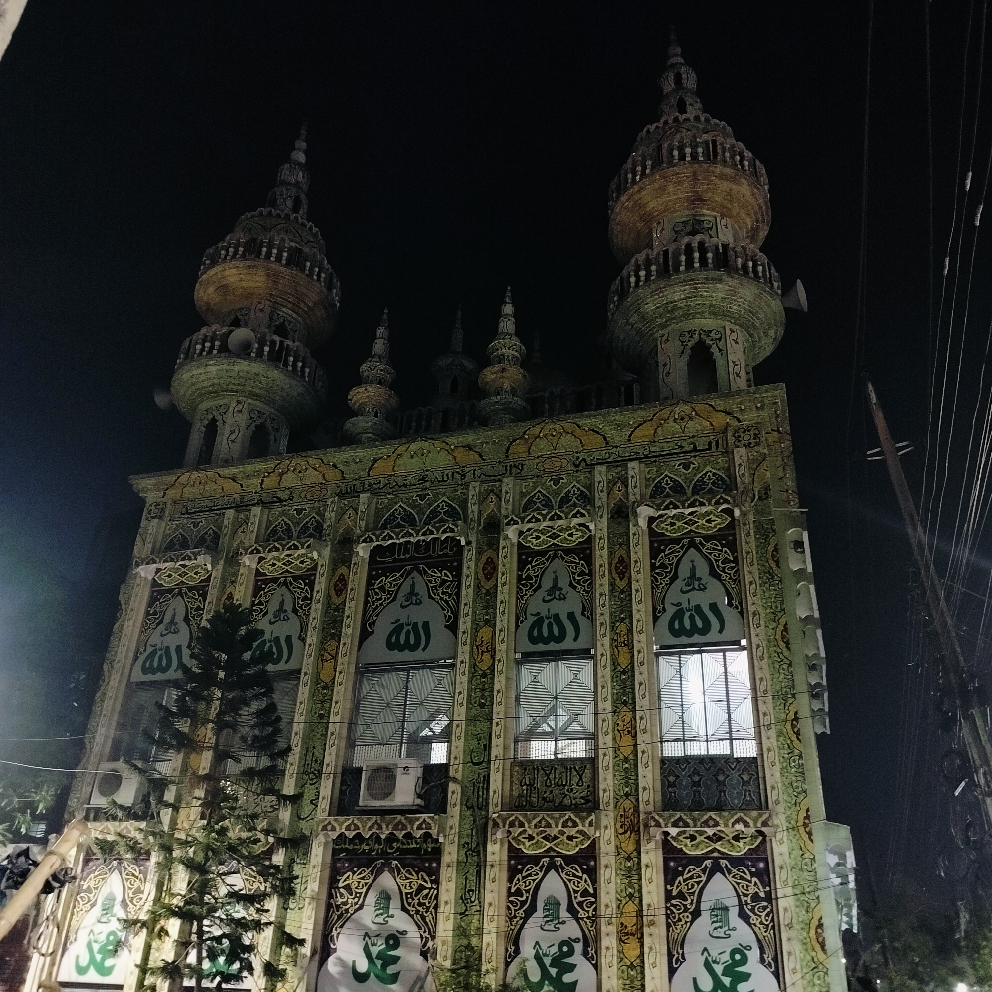 An ornate mosque stands brightly illuminated against a dark night sky in Dhaka, Bangladesh. The building's facade is richly decorated with intricate patterns in shades of green, gold, and dark blue. Two tall, multi-tiered minarets with pointed finials flank the main structure, topped with speakers for the call to prayer. Several smaller, similar spires adorn the roofline between the main minarets.
The mosque's front features multiple panels, each containing Islamic calligraphy. Prominently displayed are the names "Allah" (الله) in white script on the upper sections of the facade and "Muhammad" (محمد) in green script on white banners in the lower sections. Other intricate Arabic script, including what appears to be parts of the Shahada, is integrated into horizontal bands of the facade's design.
The windows are covered with patterned security grilles, and air conditioning units are visible below some of the upper windows. A tall, thin conifer tree partially obstructs the view of the lower left facade. To the right, utility poles and numerous electrical wires are visible, indicating an urban setting. The time of day is clearly night, with the building's lights creating a striking contrast against the dark background. No people or specific activities are depicted; the image focuses solely on the architecture and its nighttime illumination. The weather appears clear and calm.