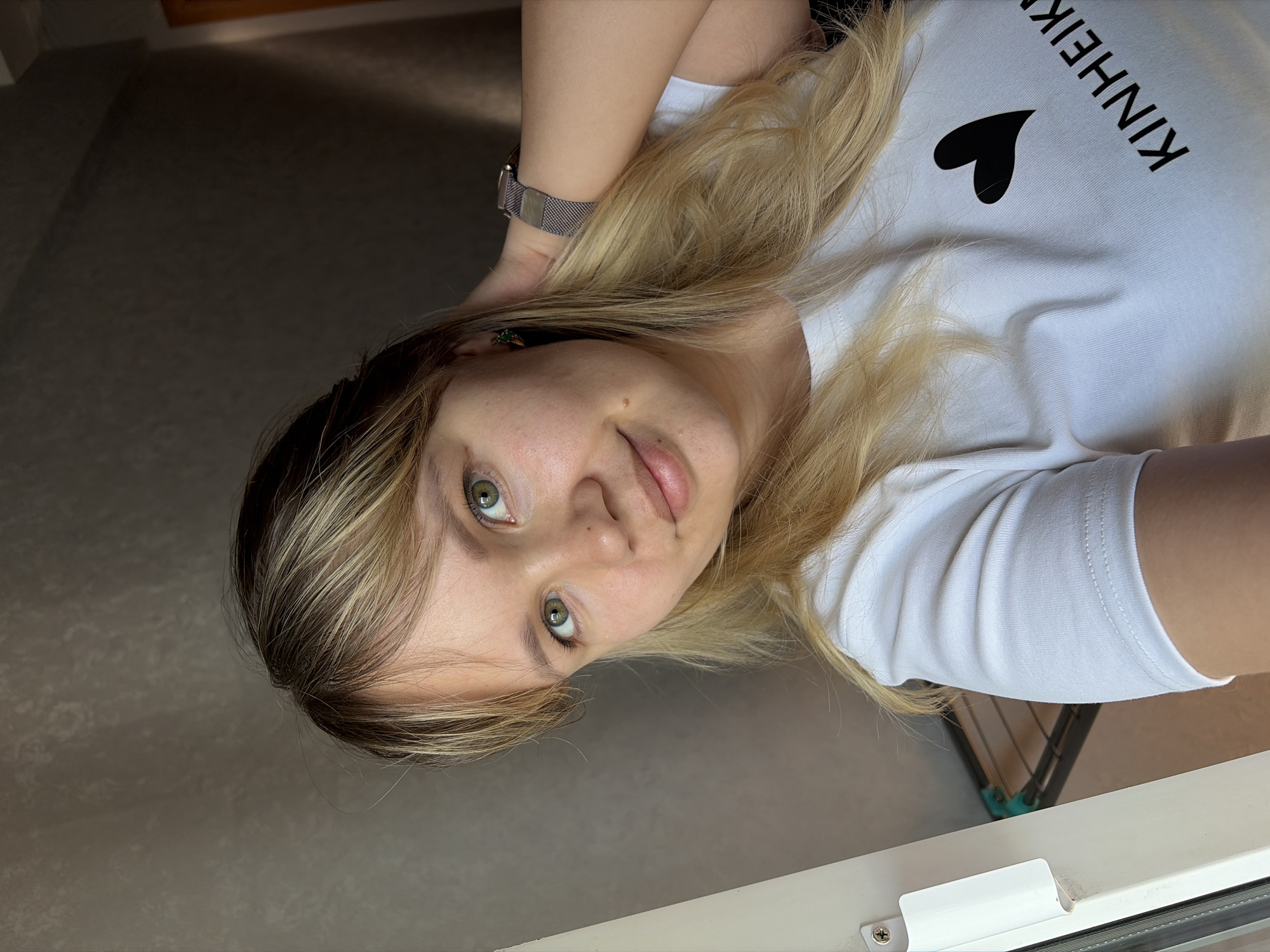 A woman with blonde hair and light skin is looking into the camera. She is wearing a white t-shirt with a black heart and the text "KINHEIK" printed on it. Her left hand, adorned with a watch featuring a metal band, is positioned behind her head. Her hair has both blonde and dark brown/black shades and is styled to the side, with a possible hair accessory visible near her left ear. The background is a mix of grey wall and white window frame. The photograph seems to be taken at an angle, possibly from above. There is no explicit information to determine the location in the city of Chaikovsky, Russia.