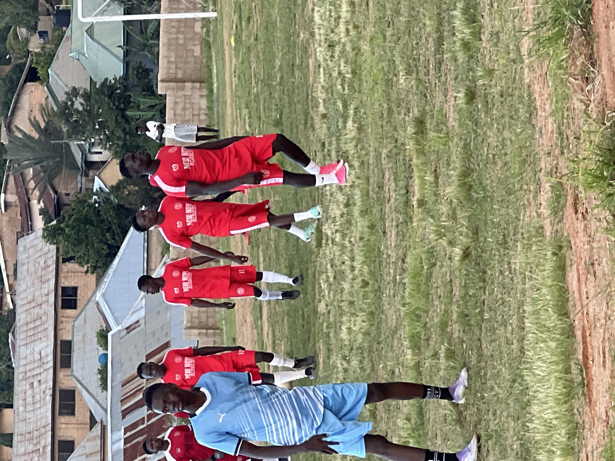 The image depicts a group of young men in athletic attire, likely a soccer team, on a grassy field. The men are predominantly wearing red jerseys and shorts, with some in blue. The setting appears to be an outdoor sports facility in Jos, Nigeria, indicated by the architectural style of the buildings in the background, typical of residential areas in the city. The buildings feature corrugated iron roofs, common in Jos due to cost-effectiveness and durability against the local climate. The field itself is a mix of green grass and reddish-brown earth, suggesting it may be a local training ground or a less-maintained stadium. In the distance, part of a soccer goal is visible, further confirming the sports context. The overall atmosphere seems casual, possibly during a practice or warm-up session.