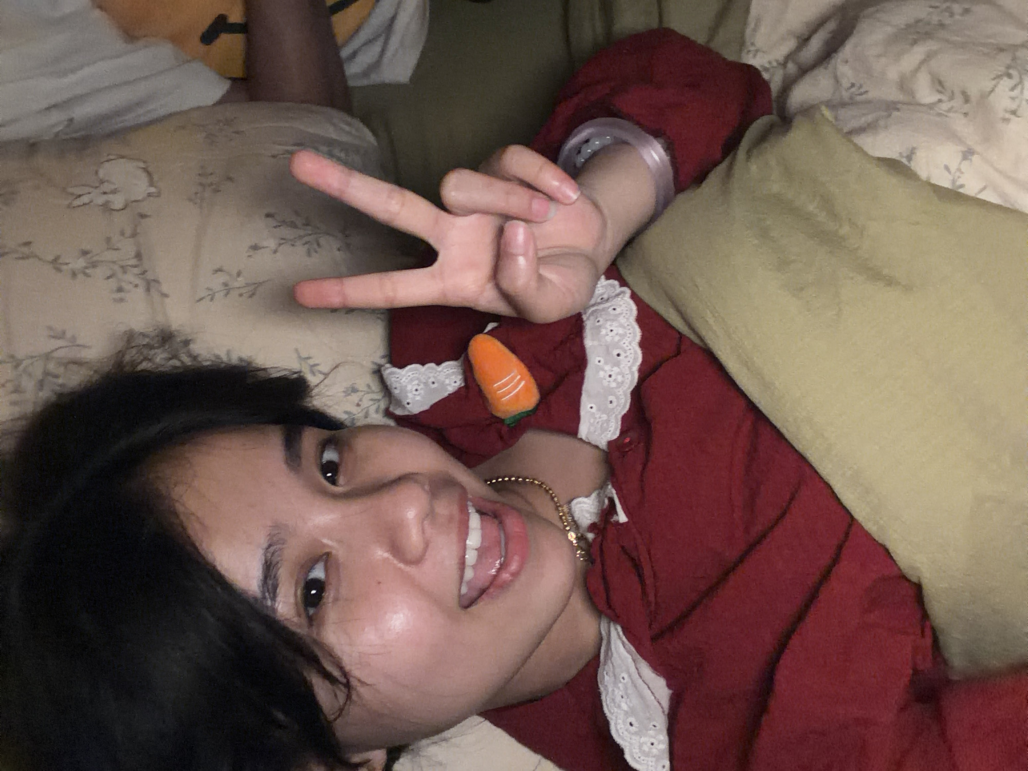An individual, likely a young female, is captured in a close-up, top-down perspective, lying down indoors, possibly on a bed or sofa. She has dark hair and a light complexion, looking directly at the camera with an open-mouthed smile, revealing white teeth and a relaxed tongue. Her eyes are dark and expressive.
She is wearing a dark red long-sleeved top featuring a white lace trim along what appears to be a collar or placket area. A thin gold chain necklace is visible around her neck, and a clear or translucent bracelet is on her right wrist. Her right hand is raised, making a peace sign (V-sign) with her index and middle fingers extended. Tucked into her clothing, near her chest, is a small, soft, orange carrot-shaped toy with white stripes and a green top, adding a playful element.
The setting appears to be a cozy indoor space, with various bedding items forming the background. These include a light-colored pillow with a subtle floral or leaf pattern, a plain light green or khaki blanket/pillow, and a white sheet or pillow. The lighting is soft and even, suggesting an indoor environment without strong natural light. The overall mood conveyed is cheerful and relaxed. There is no visible text in the image. The location context for this image is Măng Đen, Vietnam.