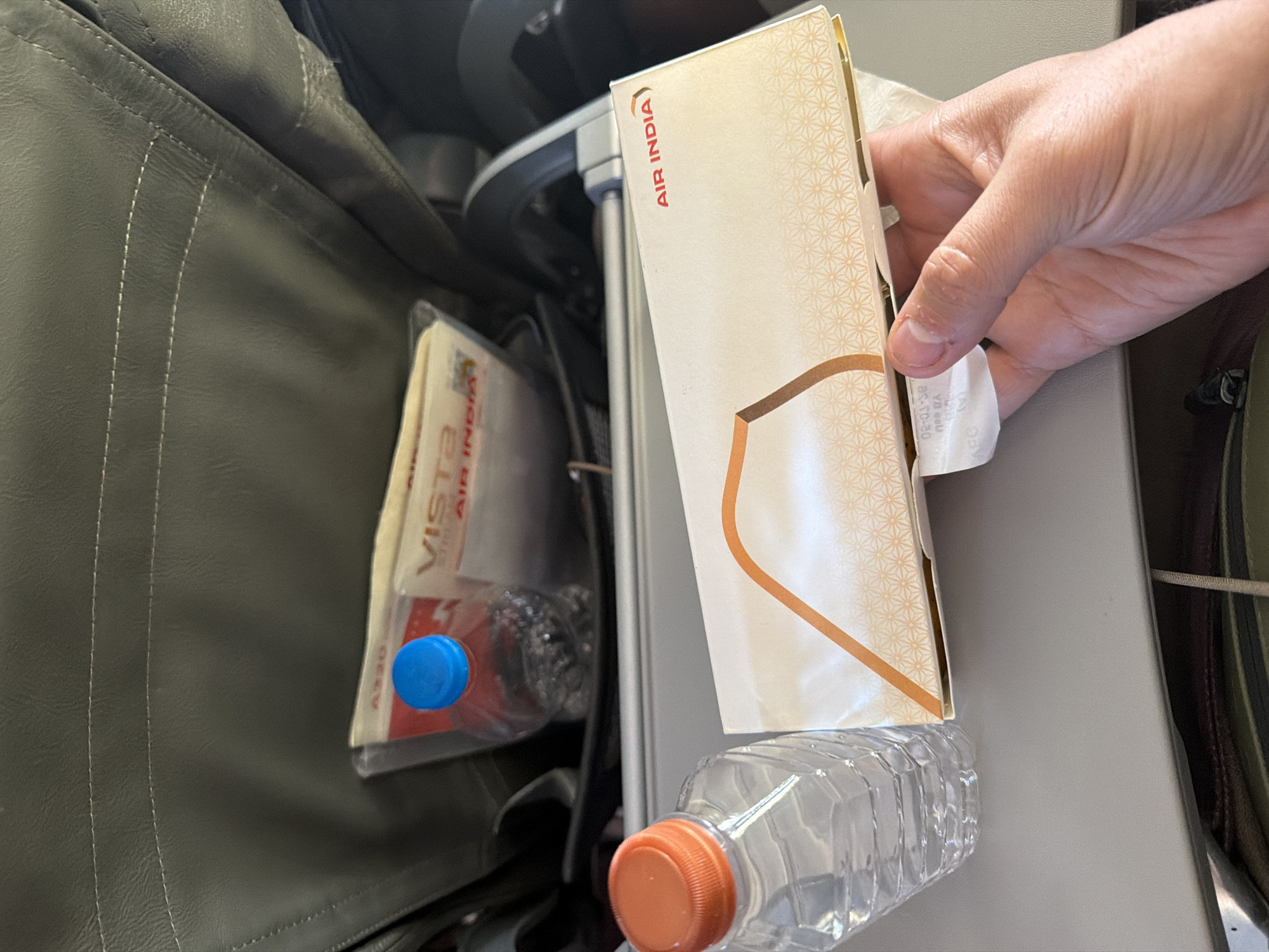The image shows a close-up view of a tray table on an airplane, likely during a flight. On the tray table, there is a box of snacks labeled "AIR INDIA" and a bottle of water with an orange cap. Behind the tray table, in the seat pocket of the airplane, there are two more water bottles, one with a blue cap, and some reading material. A hand is visible in the foreground, holding the "AIR INDIA" snack box. The overall scene suggests an in-flight meal service. There is no indication in the image that this scene is located in the city of Leh, India.