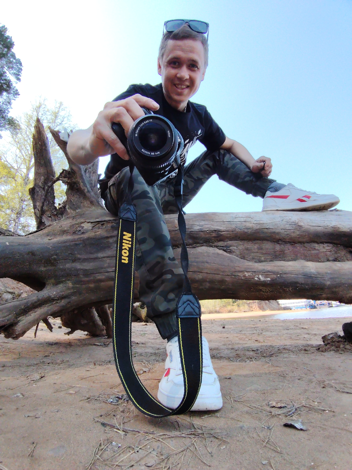 The image depicts a man holding a Nikon camera, seated on a fallen log. He is dressed in a black t-shirt and camouflage pants, and wears sunglasses atop his head. His shoes are white with red accents. The camera strap is black with the "Nikon" logo in yellow. The man is smiling and pointing the camera towards the viewer. The location appears to be a sandy area near a body of water, possibly a river or lake, with trees in the background. The scene is likely near the city of Chaikovsky, Russia, as specified in the prompt. In the background there is a building and several ships. The man's right leg is placed in front and the other leg is stretched out onto the log.