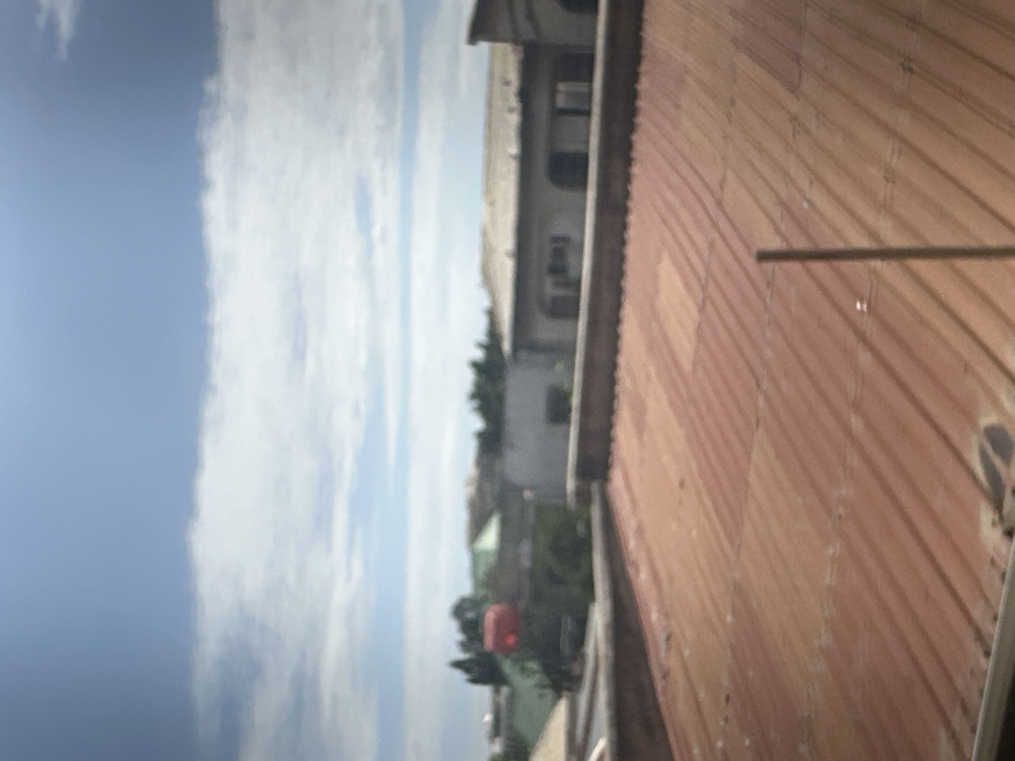The image presents an elevated, soft-focus view across a cityscape or townscape under a partly cloudy sky during daylight.
In the foreground, a weathered, reddish-brown corrugated metal roof occupies the lower half of the frame. The roof exhibits color variations and discolored patches, consistent with age or exposure. A thin, vertical, light-colored rod extends upwards from the bottom edge, positioned slightly right of center.
Immediately beyond the foreground roof, a dark, horizontal parapet wall spans the width of the image. Behind this wall, various structures are visible. To the left, a building with a prominent red cylindrical tank on its roof is partially obscured by green foliage. To the right, a lighter-colored building with a flat, light-toned roof displays multiple arched openings or recesses along its visible facade.
The background consists of indistinct structures and sparse vegetation. The upper portion of the image is dominated by a light blue sky, featuring a prominent, horizontal band of white clouds across its mid-section, with additional scattered clouds visible.
The entire scene is captured with a general softness of focus, hindering precise identification of distant details. No individuals are present in the frame, and no specific actions or interactions are discernible. The location is an unknown city, unknown.