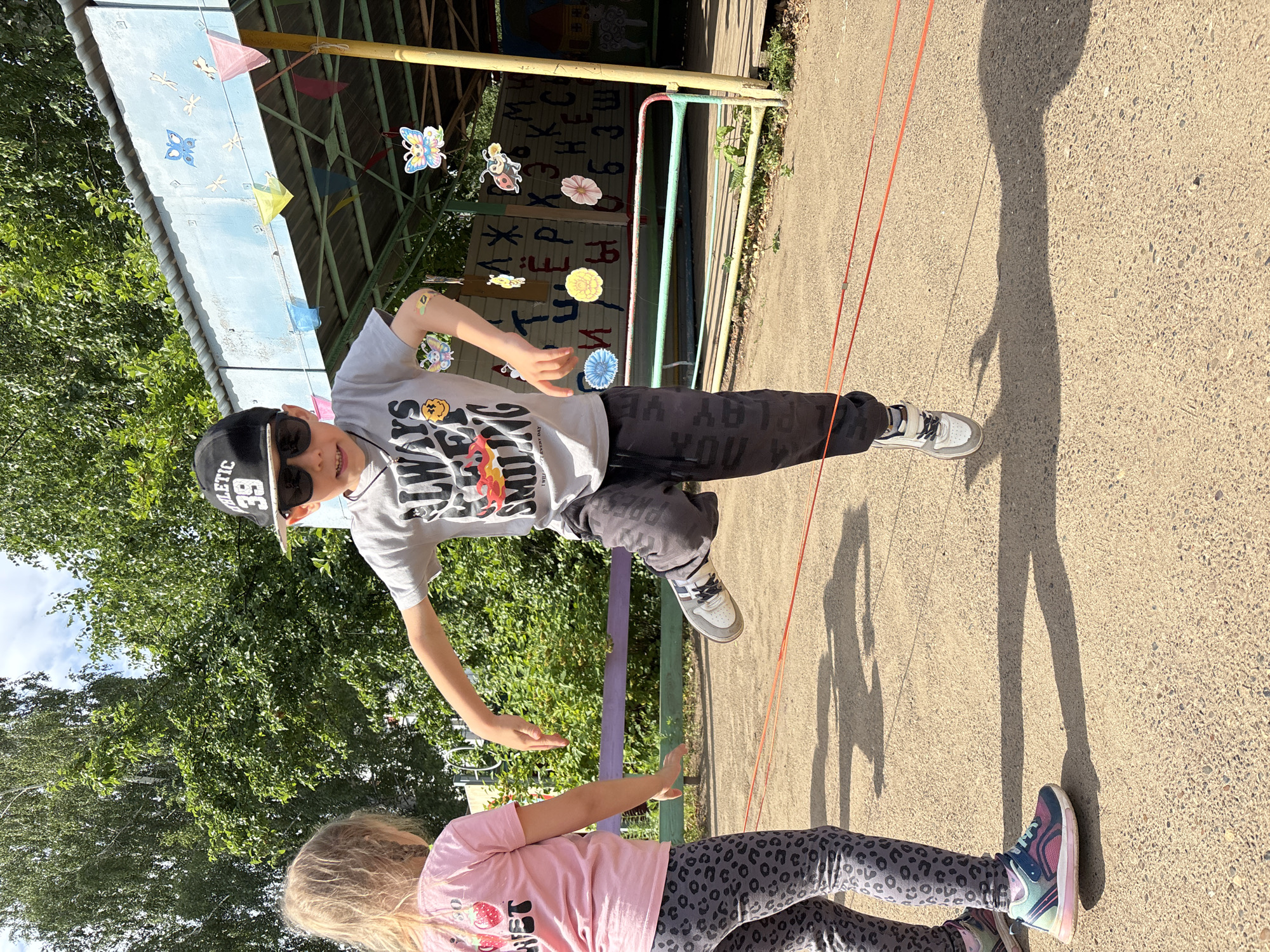 The image depicts two children, a boy and a girl, engaged in a game of jump rope outdoors. The boy, wearing a black baseball cap with the number "39" and sunglasses, is mid-jump. He sports a grey t-shirt with the text "ALWAYS SMILING" and dark pants. The girl, with blonde hair, is wearing a pink t-shirt with a strawberry graphic and grey leopard-print leggings. Both children are barefoot or wearing light-colored sneakers. The scene appears to be a playground or an outdoor recreational area, possibly a schoolyard, given the presence of a covered structure with colorful decorations like butterflies and flowers, and what looks like letters painted on a wall in the background. The ground is paved with a light-colored, grainy surface, and shadows indicate it is daytime with bright sunlight. The overall atmosphere seems cheerful and active. Visible text includes "ATHLETIC 39", "ALWAYS SMILING", and "YOU". The location is identified as Chaikovsky, Russia.