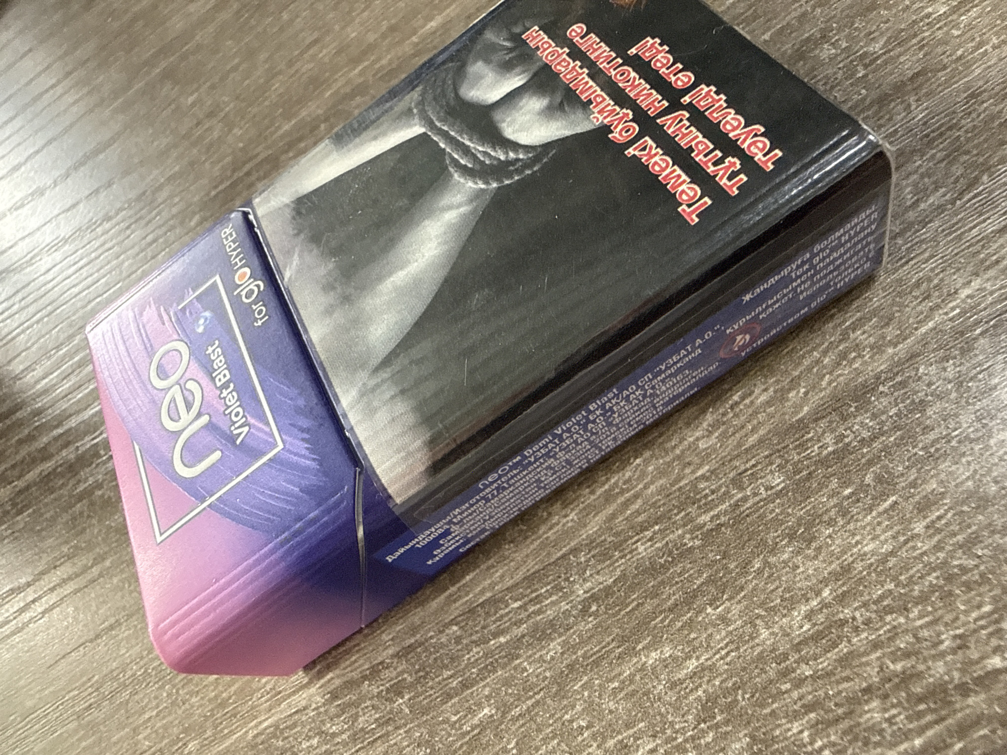 The image shows a pack of Neo Demi Violet Blast tobacco sticks for use with a glo HYPER device. The pack is placed on a wooden surface. The front of the pack features the "Neo" logo in large, stylized white lettering within a geometric shape, transitioning from pink to purple. To the left of the logo, it states "Violet Blast" and "for glo HYPER" in smaller text. The top portion of the pack displays an image of hands bound by rope, with text in both Kazakh and Russian. The Kazakh text reads "Темекі бұйымдарын тұтыну никотинге тәуелді етеді" which translates to "Tobacco products cause nicotine addiction." The Russian text conveys a similar warning. Below this, there is information in Russian, including "Дайындаушы/Изготовитель: "УЗБАТ А.О." БК АК/АО СП "УЗБАТ А.О.", Өзбекстан/Узбекистан" indicating the manufacturer. There are also mentions of manufacturing dates and product codes. The overall impression is a product package designed to appeal to consumers while also adhering to regulatory requirements regarding health warnings. The lighting suggests an indoor setting, possibly a table, with no clear indication of time of day or weather. The scene is focused on the product itself.