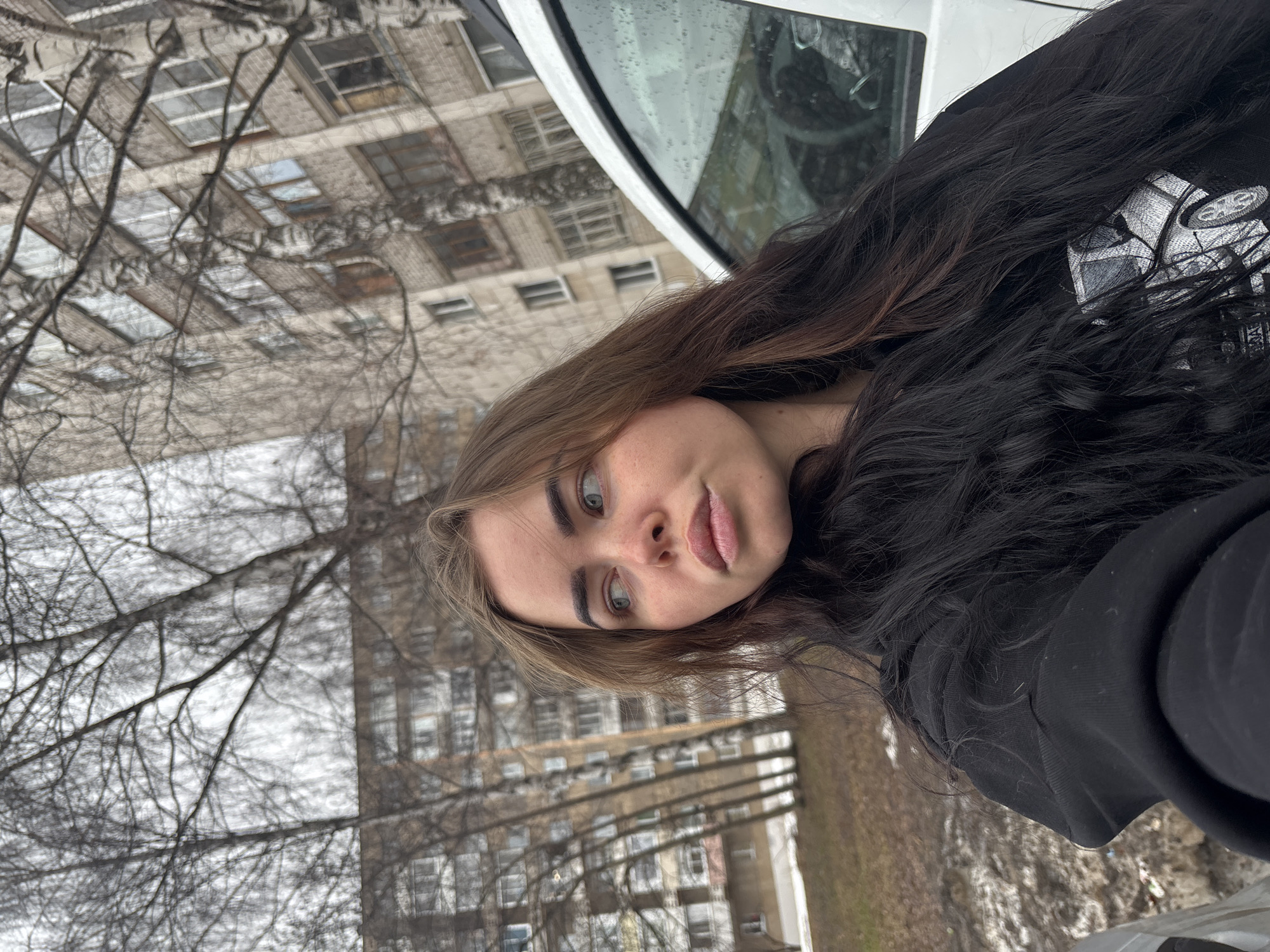 A young woman with light skin, light eyes, and dark and brown long hair takes a selfie outdoors. She wears a black hooded sweatshirt. Behind her are bare trees, a grassy area with patches of snow, and a multi-story building. Part of a white vehicle is visible at the top of the frame. This image appears to have been taken in Izhevsk, Russia.