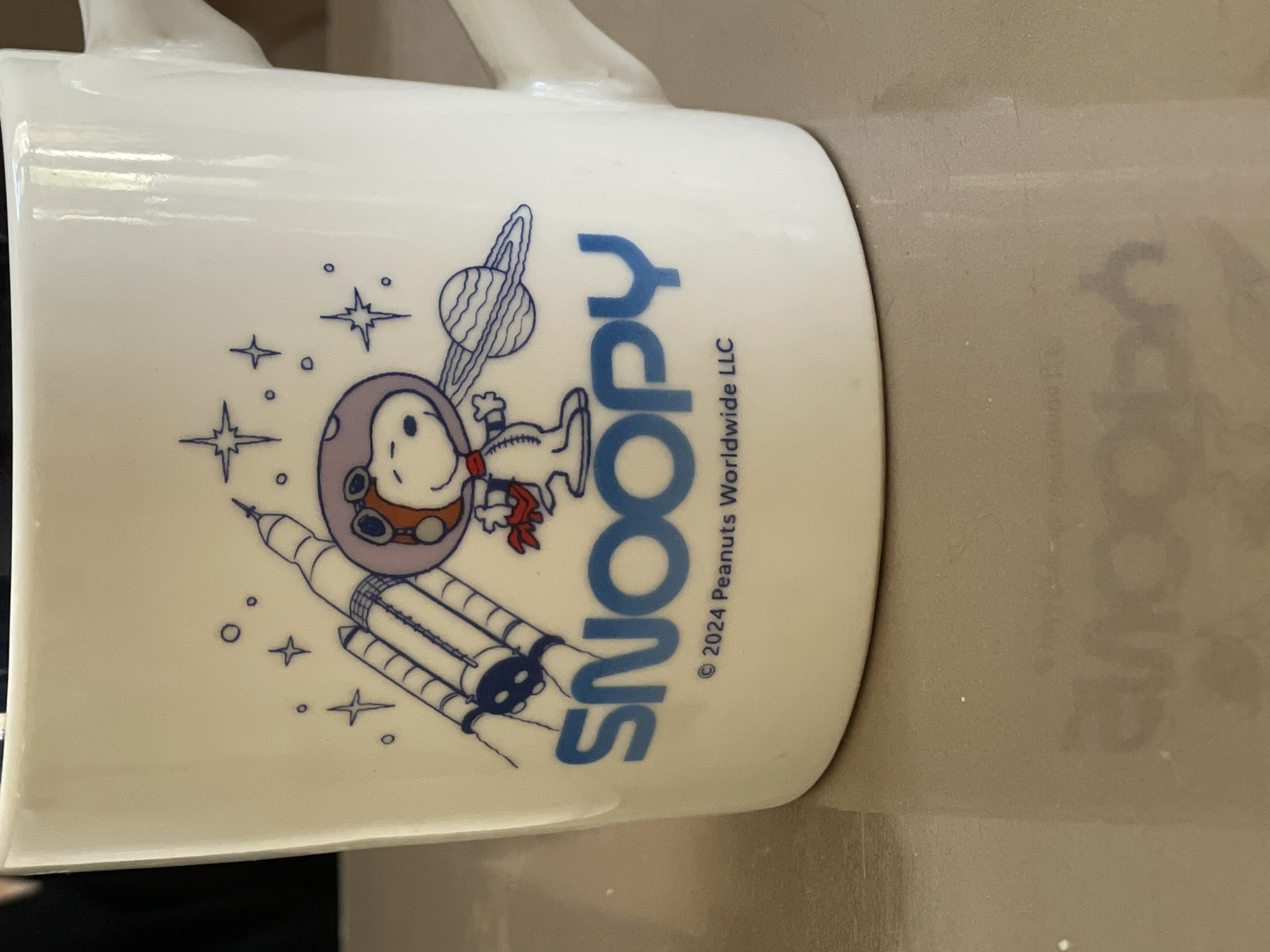 A white ceramic mug with blue graphics is positioned on a reflective brown surface. The mug features an illustration of Snoopy in a space suit, a rocket, and planets, with the word "SNOOPY" printed in large blue letters. Below the name, the text "© 2024 Peanuts Worldwide LLC" is present. The background is blurred, but appears to be a dark interior. The image is a close-up of the mug, with no visible elements indicating a specific location in Tashkent, Uzbekistan.