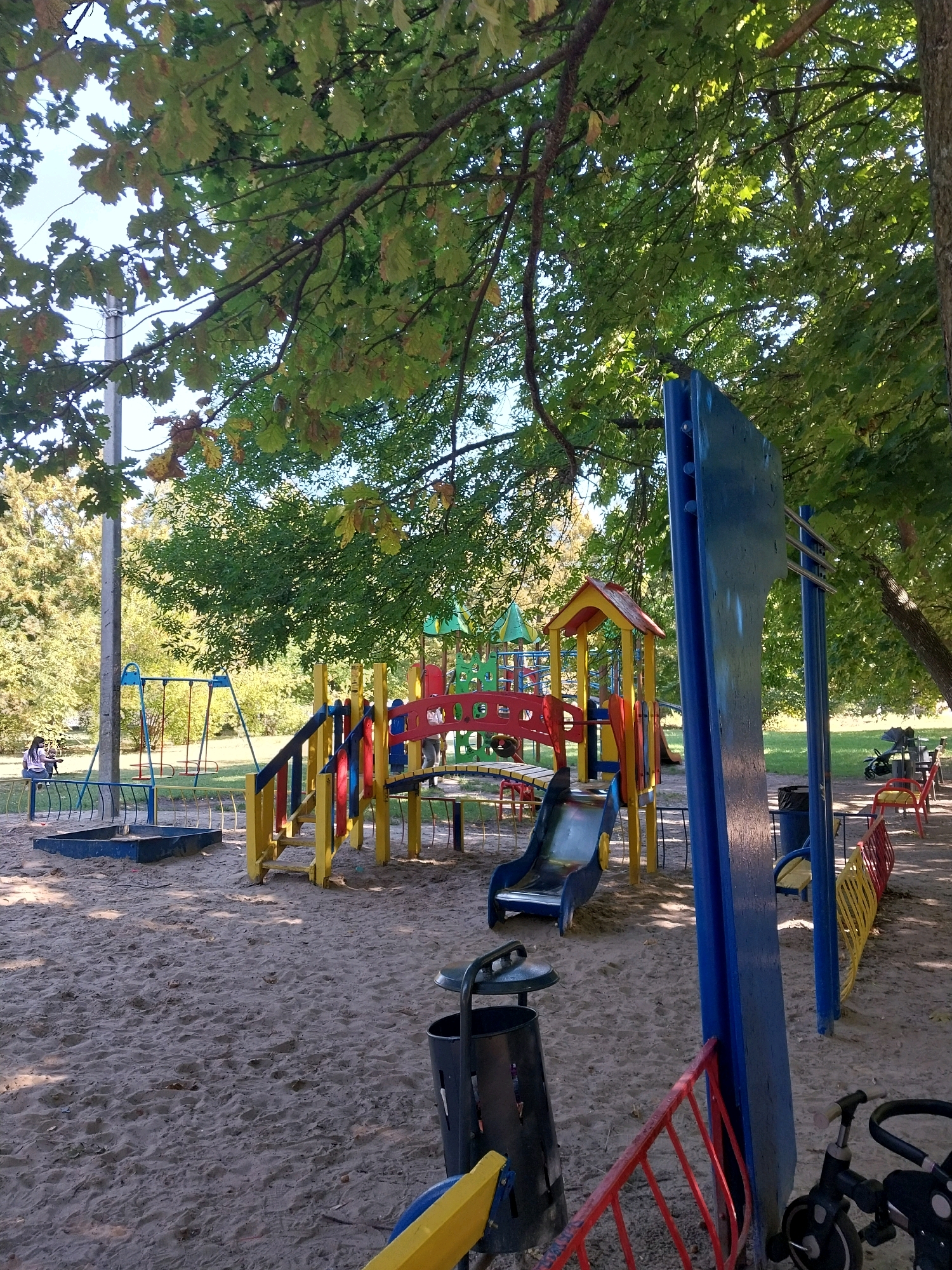 The image depicts a children's playground situated in the city of Doslidne, Ukraine. The playground is constructed on a sandy surface, surrounded by trees providing ample shade. Key features of the playground include a blue sandbox, a swing set with red seats, a multi-colored climbing structure with yellow and blue supports and red and blue railings, and a blue slide. Adjacent to the climbing structure, a blue and yellow bench is visible. Further into the background, a yellow and red segmented fence lines a grassy area. A black trash can is positioned in the foreground. In the distance, a black children's tricycle is parked. The presence of autumn leaves on the trees suggests the season is likely fall.