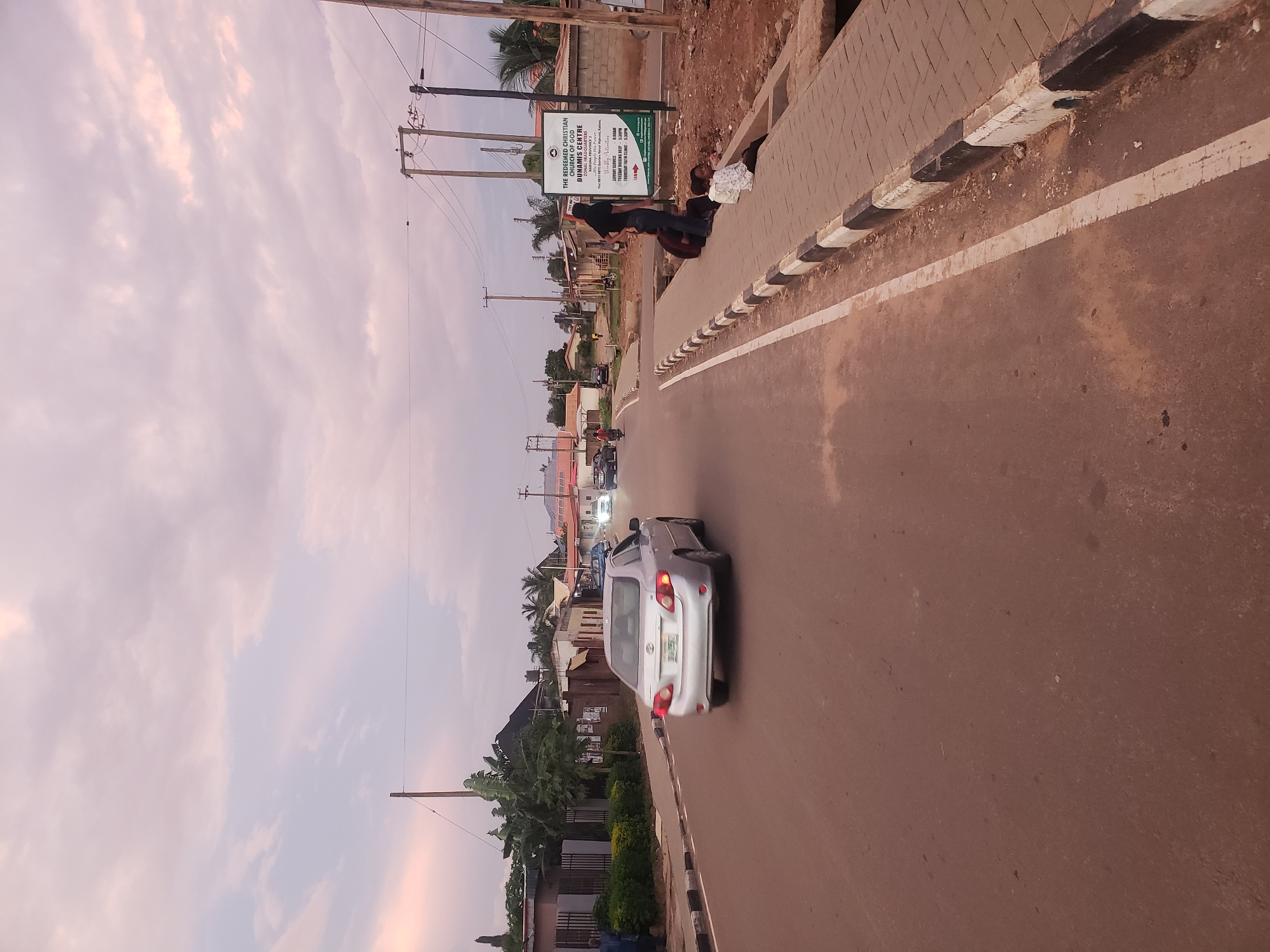 The image depicts a street scene in Narayi, Nigeria. A silver sedan is driving down the road, with a white line marking the lane. On the right side of the road, a sidewalk is lined with black and white bollards. Several people are sitting on the edge of the sidewalk, and a large sign for "THE REDEEMED CHRISTIAN CHURCH OF GOD DUNAMIS CENTRE" is visible behind them. The sky is overcast with pink and grey clouds. Buildings and trees are visible in the background.