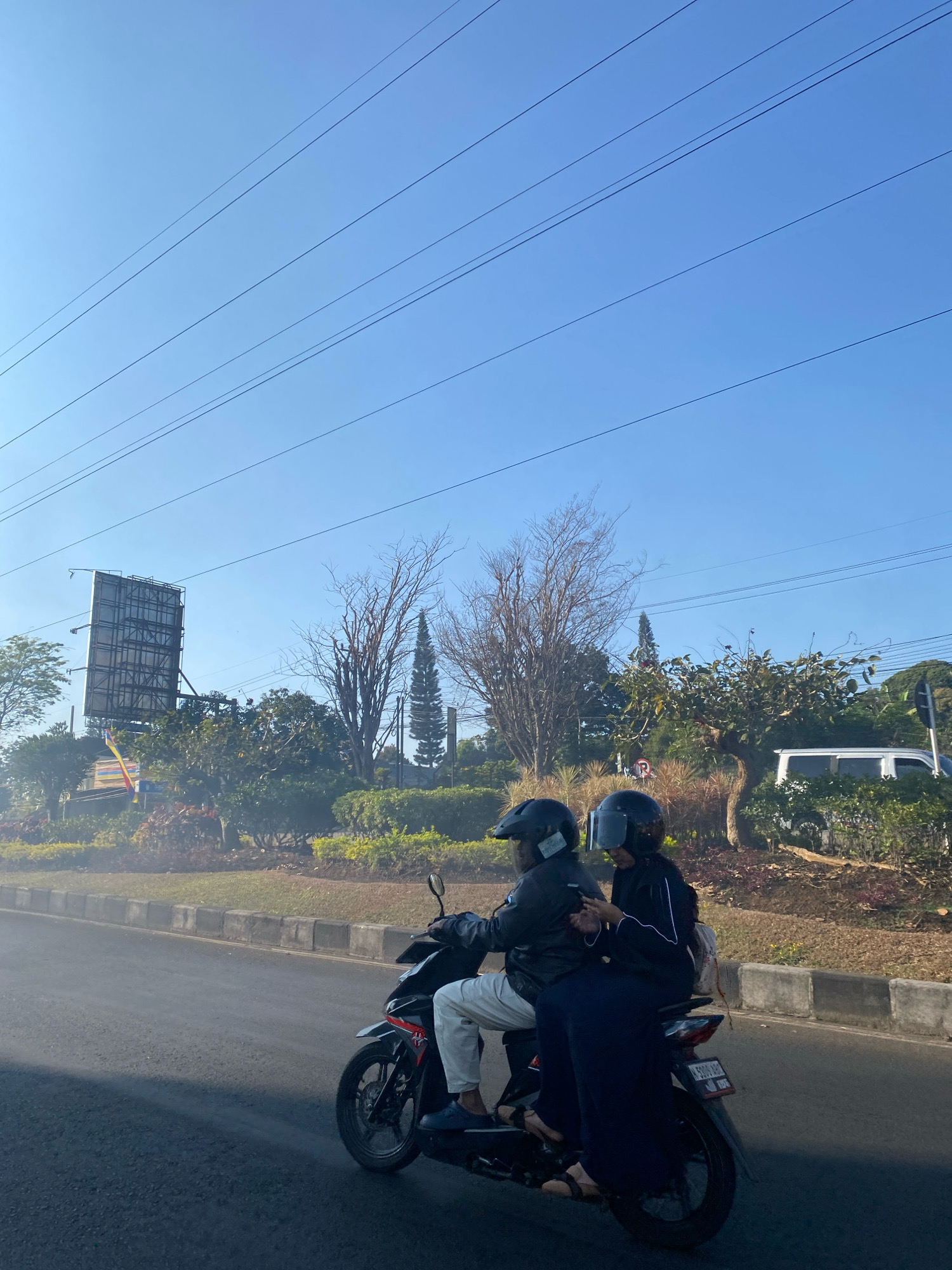 Two individuals, wearing helmets, ride on a black motorcycle on a paved road. The person in front is wearing a black jacket and white pants, while the person behind is wearing a black garment and dark blue pants. The motorcycle is moving from left to right. In the background, there are trees, bushes, a billboard, power lines, and a white van. The sky is clear and blue.