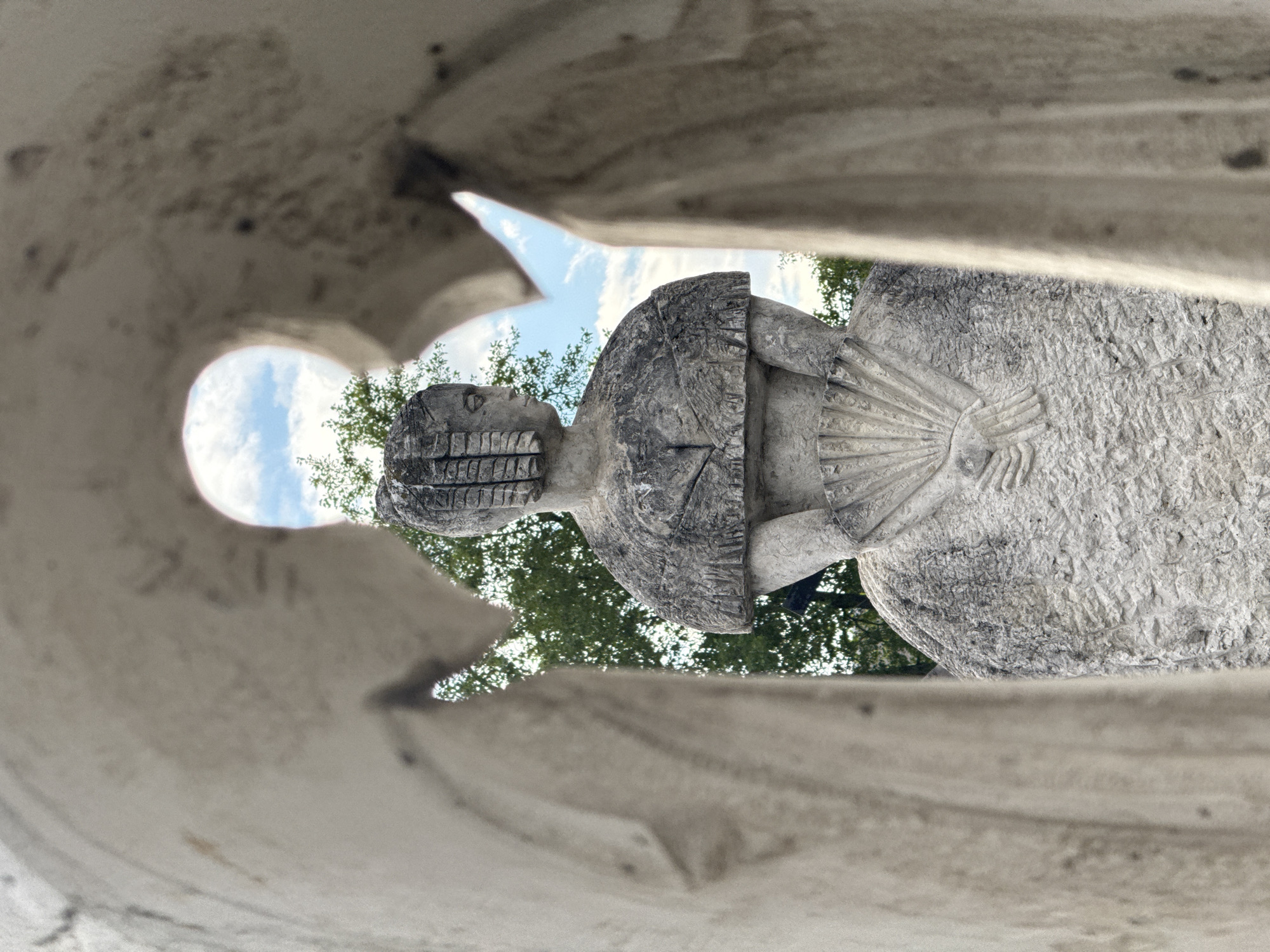 The image is a close-up, eye-level, outdoor shot of a stone sculpture of a woman. The sculpture is framed by two other stone elements, creating a window effect. The woman is depicted from the torso up, wearing a voluminous dress and a textured shawl. Her hair is styled in vertical rows, and she holds a fan in her clasped hands behind her back. The sculpture has a rough, weathered texture. Behind the sculpture, a bright blue sky with scattered white clouds is visible, along with lush green foliage from trees. The overall impression is one of an artistic display in an outdoor setting.