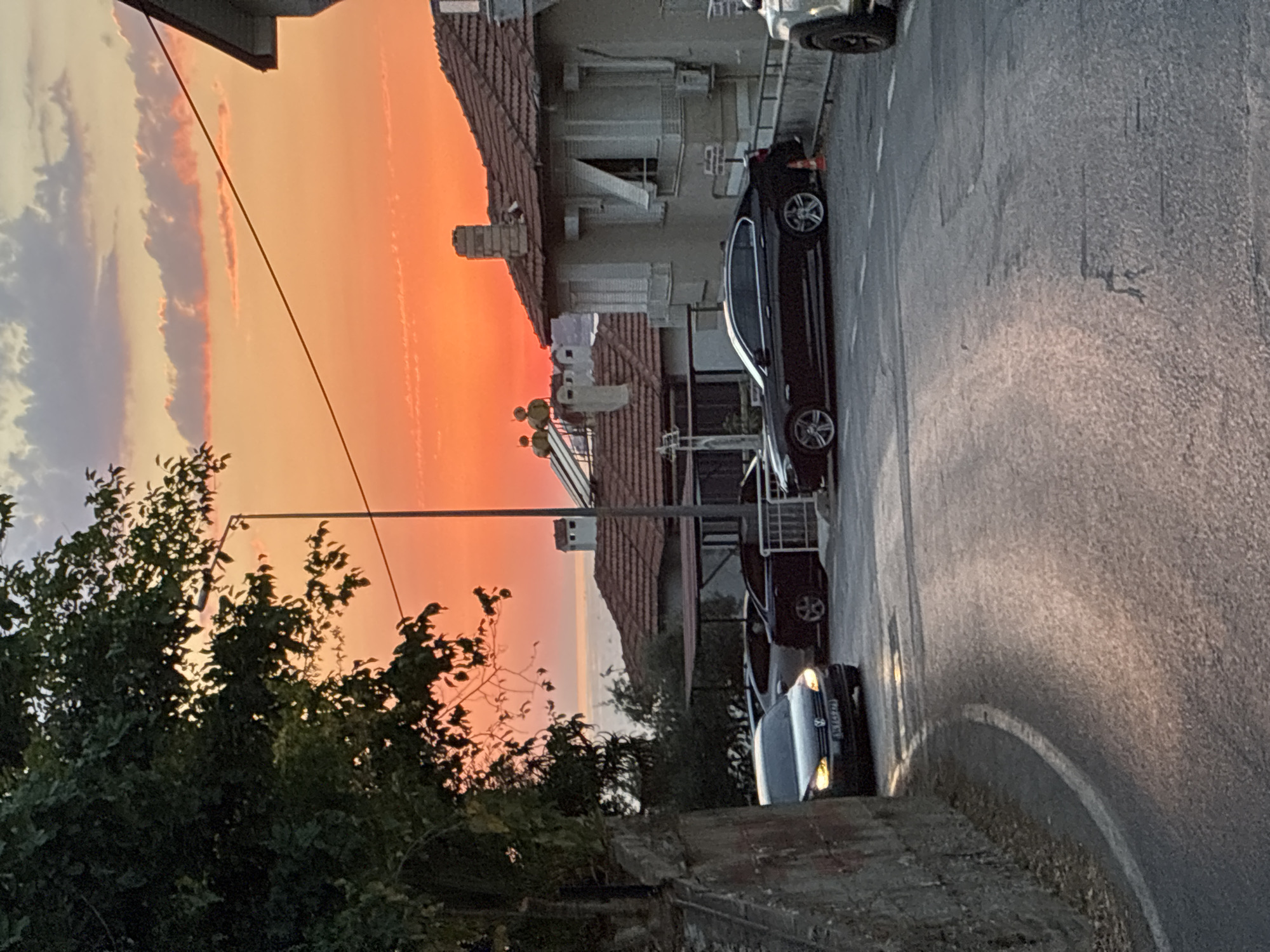 This image captures a sunset scene in a coastal town, likely Alanya, Turkey, given the architecture and the Mediterranean Sea visible in the background. The sky is a vibrant gradient of orange, pink, and yellow, with scattered clouds. A street curves into the foreground, reflecting the warm sunset light. Several cars are parked along the street, including a black sedan and a lighter-colored car partially visible at the bottom right. Residential buildings with tiled roofs and characteristic window shutters line the street. Dense foliage from a large tree on the left frames a portion of the scene. A streetlamp is also visible. The overall atmosphere is peaceful and serene, typical of a late afternoon in a Mediterranean locale.
