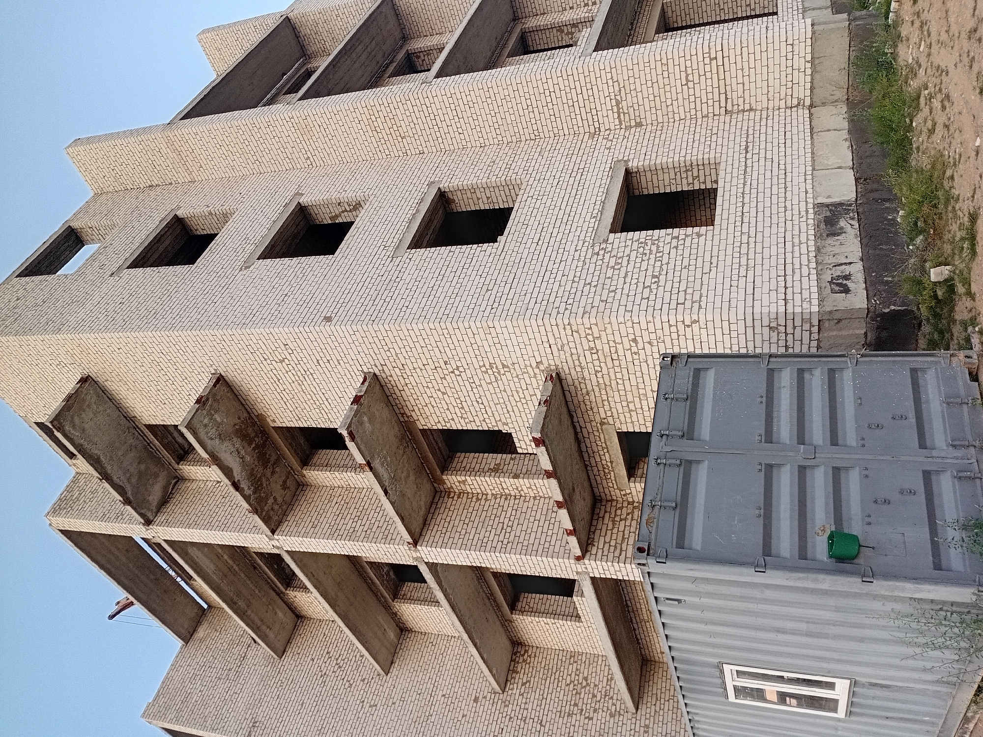 The image shows an unfinished multi-story brick building under construction in the city of Semey, Kazakhstan. The building has a light-colored brick exterior and concrete balconies or floor slabs extending from each level. Many of the window openings are empty. In the foreground, a large, grey, corrugated metal shipping container with a small white window is positioned next to the building. A green bucket is attached to the side of the container. The ground around the construction site appears to be dirt and sparse vegetation. The sky is clear and light blue, suggesting daytime. A construction crane is visible at the top of the building in the distance.