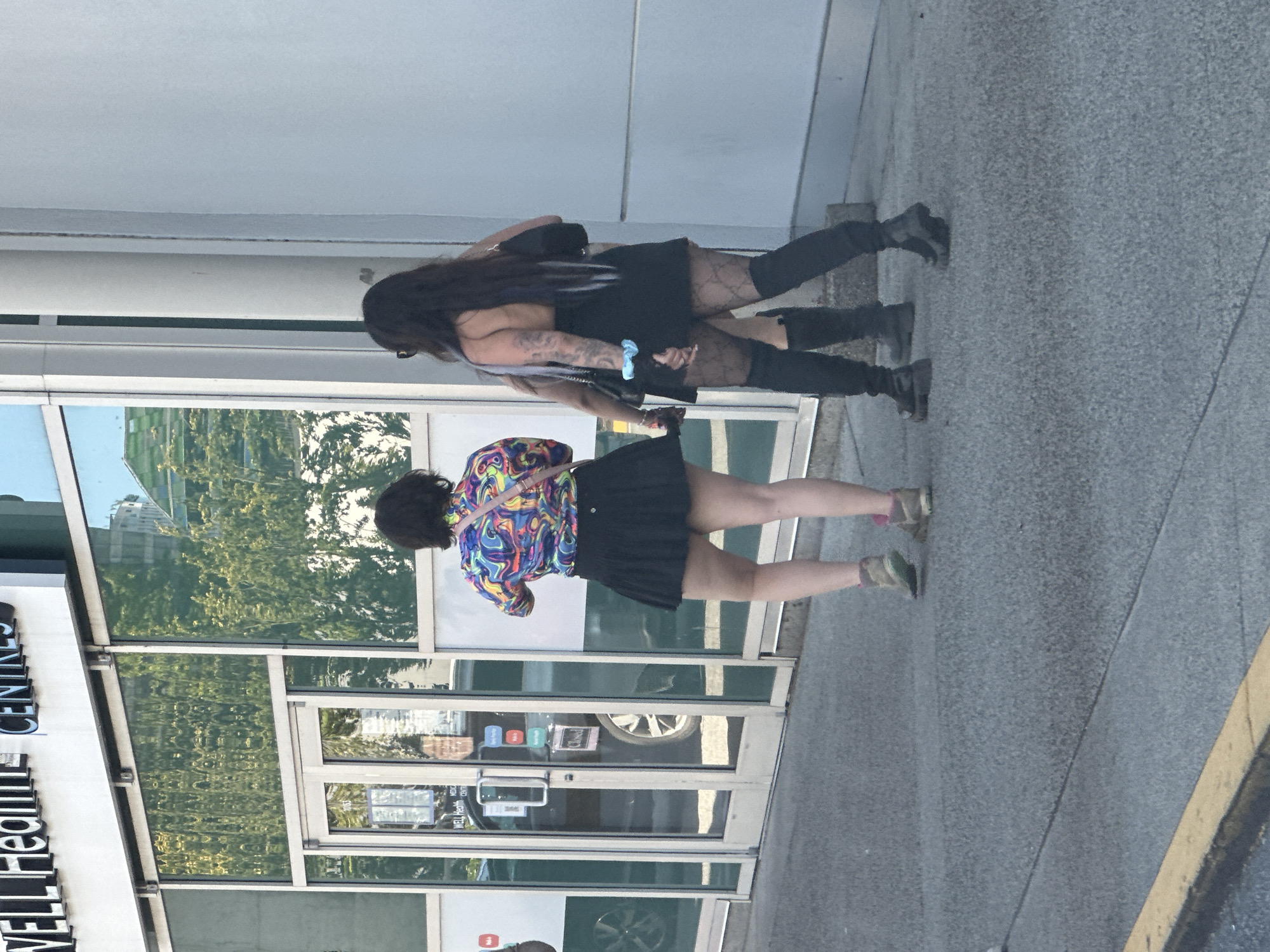 A group of people are standing outside a building with a sign that reads "WELL Health CENTRES". The building appears to be a medical facility with large glass windows reflecting trees and a building. One person is wearing a colorful patterned shirt and a black skirt. Another person is wearing a black dress and patterned tights, with a tattoo visible on their arm. The ground is paved with a light grey asphalt. A yellow curb is visible in the foreground. The image was taken outdoors on a sunny day.